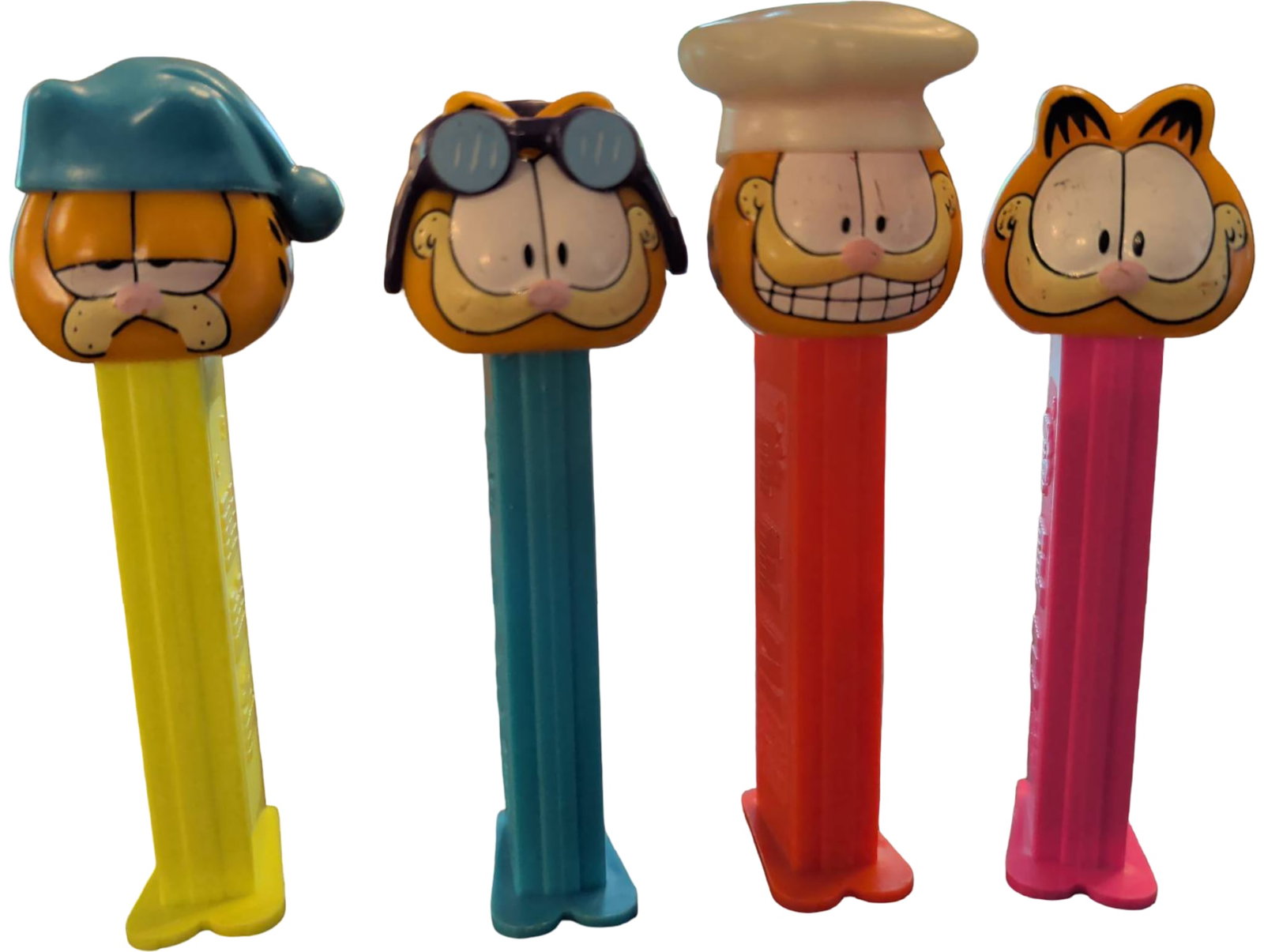 Four Garfield-Themed PEZ Dispensers: Four collectible PEZ dispensers featuring Garfield character designs. Each dispenser depicts Garfield in different outfits, including a blue sleeping cap, aviator goggles, a chef hat, and a classic lo