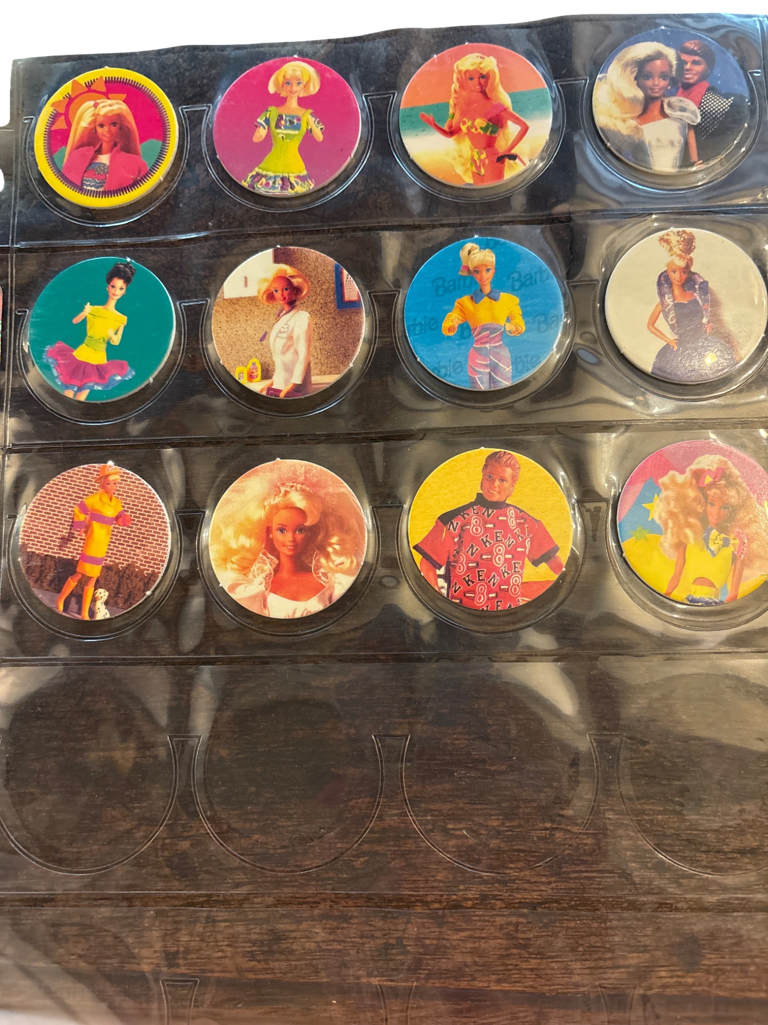 Barbie-Themed Collectible Pogs with Plastic Album Sheet: 12 Barbie-themed collectible pogs featuring various iconic Barbie designs and outfits. Each pog is circular and brightly colored, housed in a clear plastic album sheet. The sheet has individual slots 