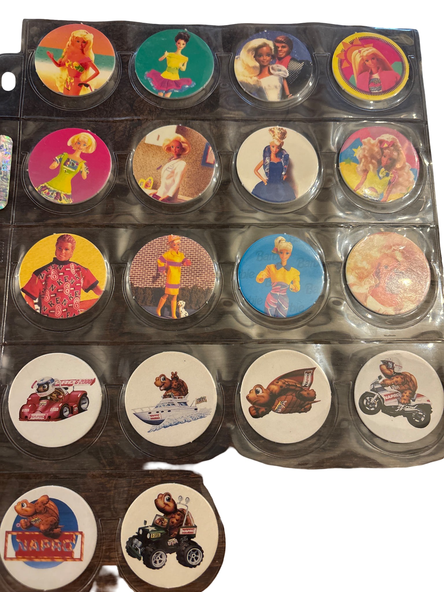 Pog Milk Caps Featuring Various Designs: 18 Pog milk caps housed in a protective plastic sleeve. Each cap features colorful designs, including characters, animals, and vehicles. Materials include printed cardboard with slight signs of handli