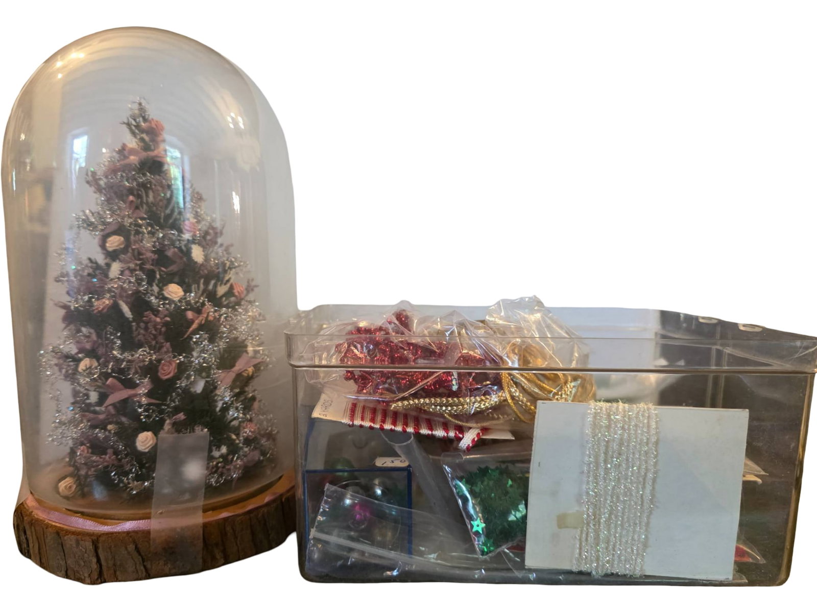 Decorative Miniature Christmas Tree in Glass Dome with Additional Christmas Decorations for: Miniature Christmas tree with pink ribbon, floral ornaments, and tinsel detail, enclosed in a glass dome with wooden base. Includes a plastic box containing assorted craft supplies such as ribbons, be