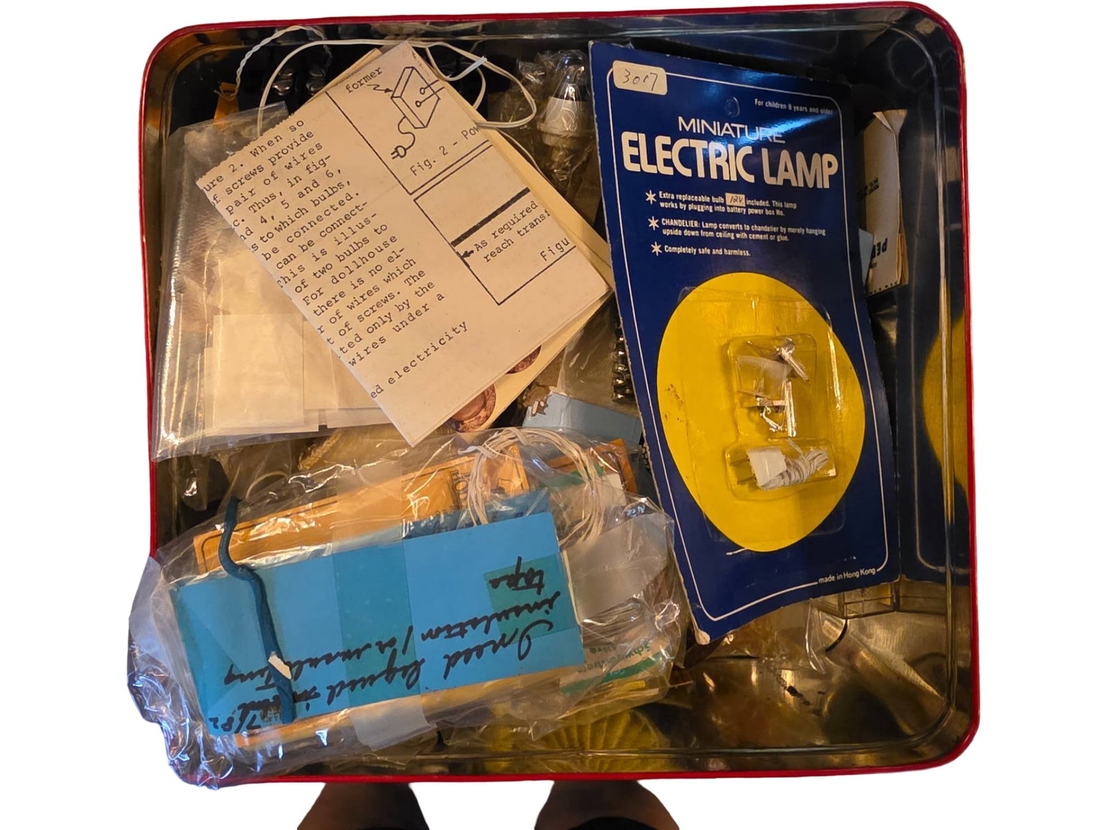 Dollhouse Electronics, Components and Accessories in a Tin Container: Small electronic components and accessories housed in a tin container. Includes items such as a packaged electrical lamp, various wires, a handwritten note, connectors, and other components. The tin c