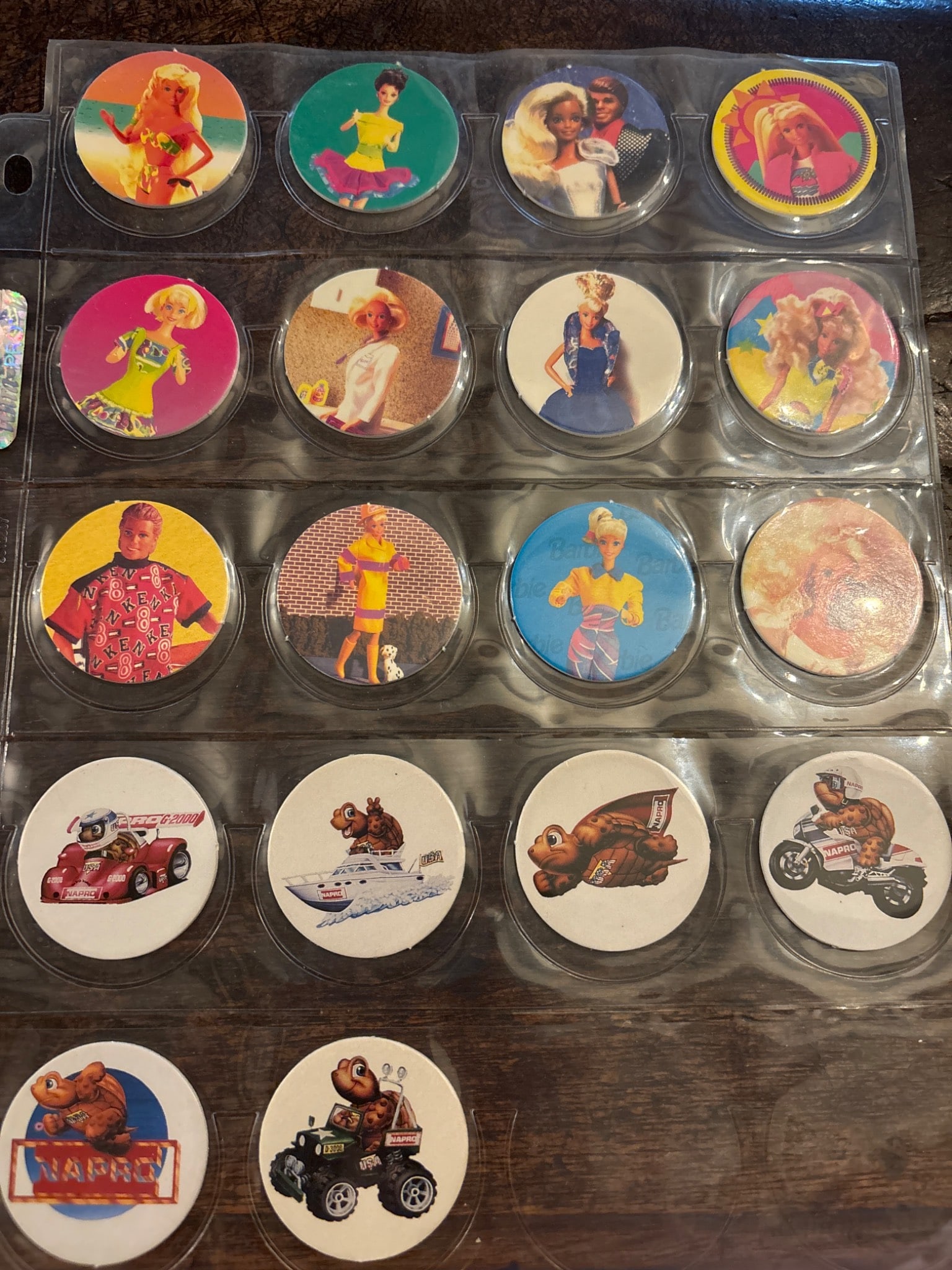 Pog Milk Caps Featuring Various Designs: 18 Pog milk caps housed in a protective plastic sleeve. Each cap features colorful designs, including characters, animals, and vehicles. Materials include printed cardboard with slight signs of handli