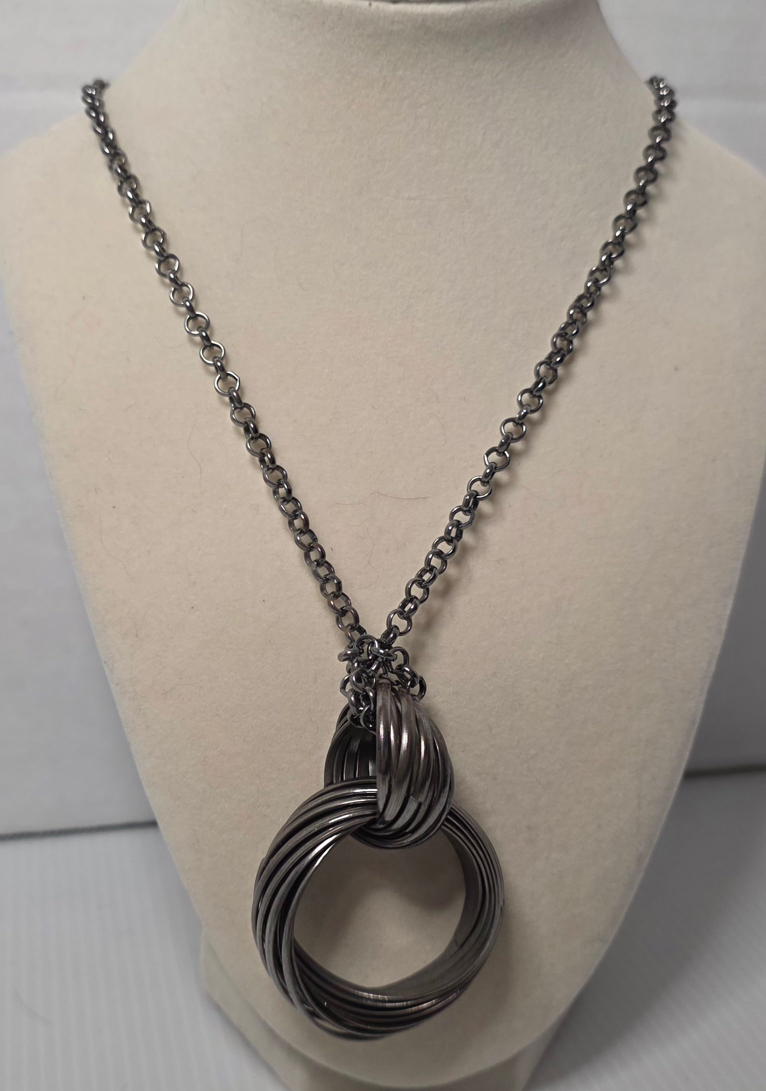Gunmetal Tone Multi Ring Pendant Statement Necklace: Gunmetal tone chain necklace featuring a large multi ring circular pendant. Interlocking stacked rings create a bold modern statement design. Lobster clasp closure. Measures approximately 28 inches ov