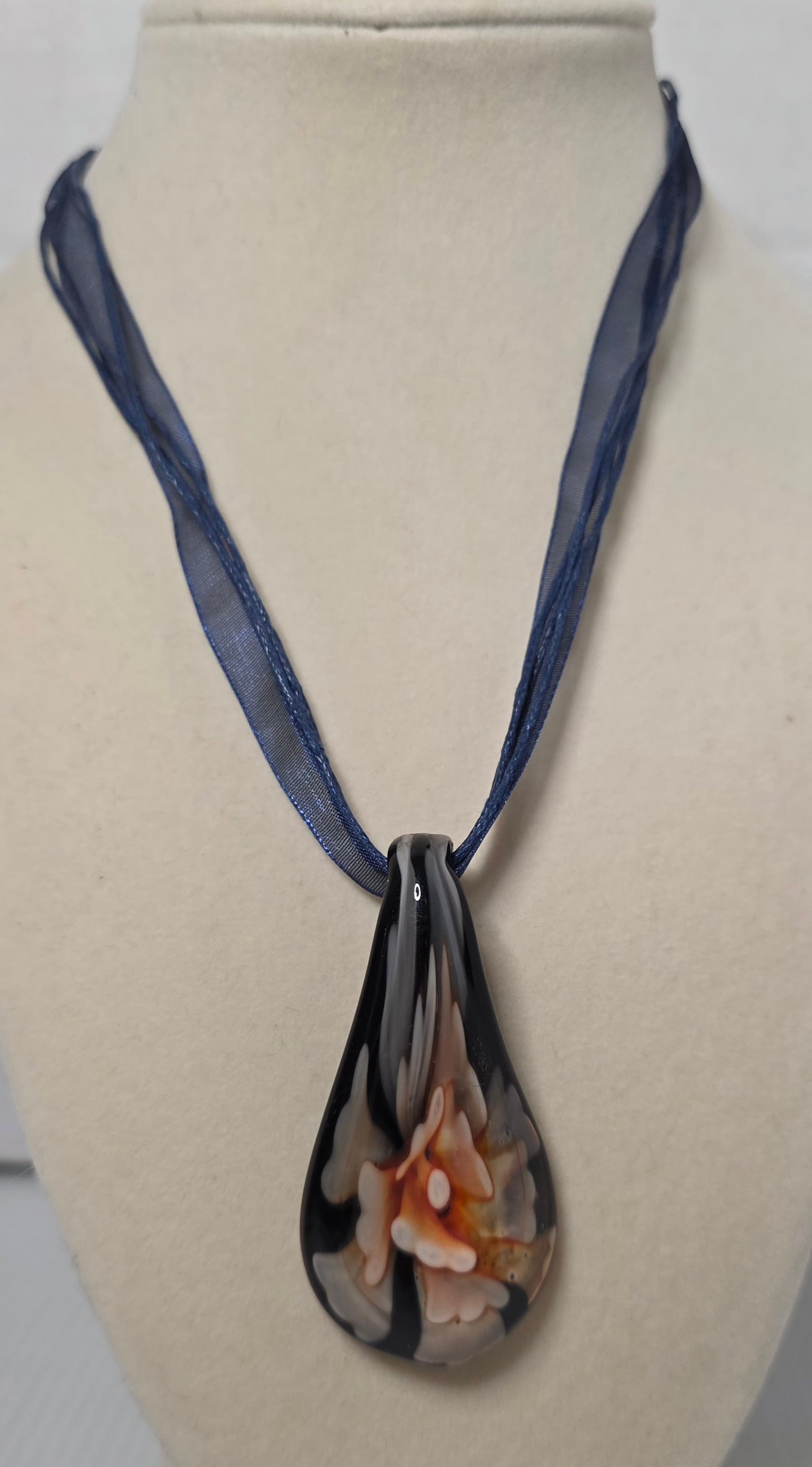 Art Glass Teardrop Pendant Blue Ribbon Necklace: Large art glass teardrop pendant featuring a raised floral design in cream and amber tones over a dark background. Suspended from layered navy blue ribbon and cord strands with lobster clasp and exten