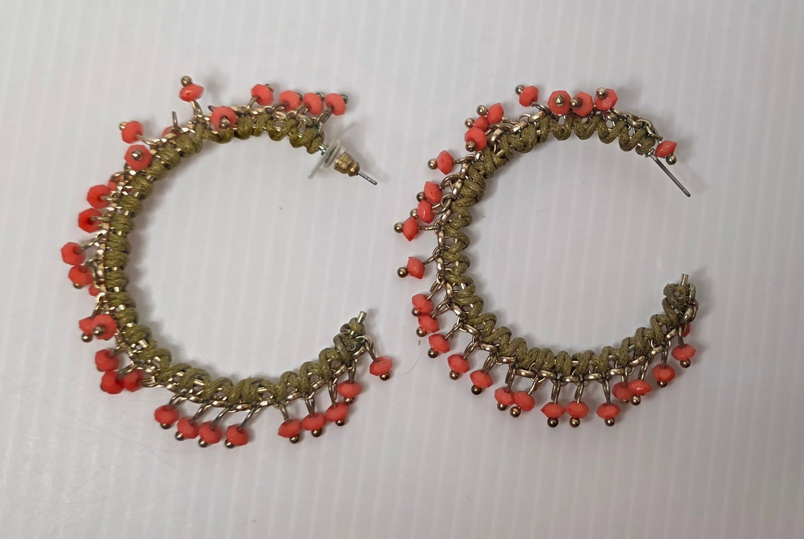 Boho Coral Bead Wrapped Hoop Earrings Gold Tone Pair: Pair of boho style hoop earrings featuring wrapped green cord over gold tone metal with dangling coral color bead accents and small gold tone ball details. Post back style.Hoops measure approximately 