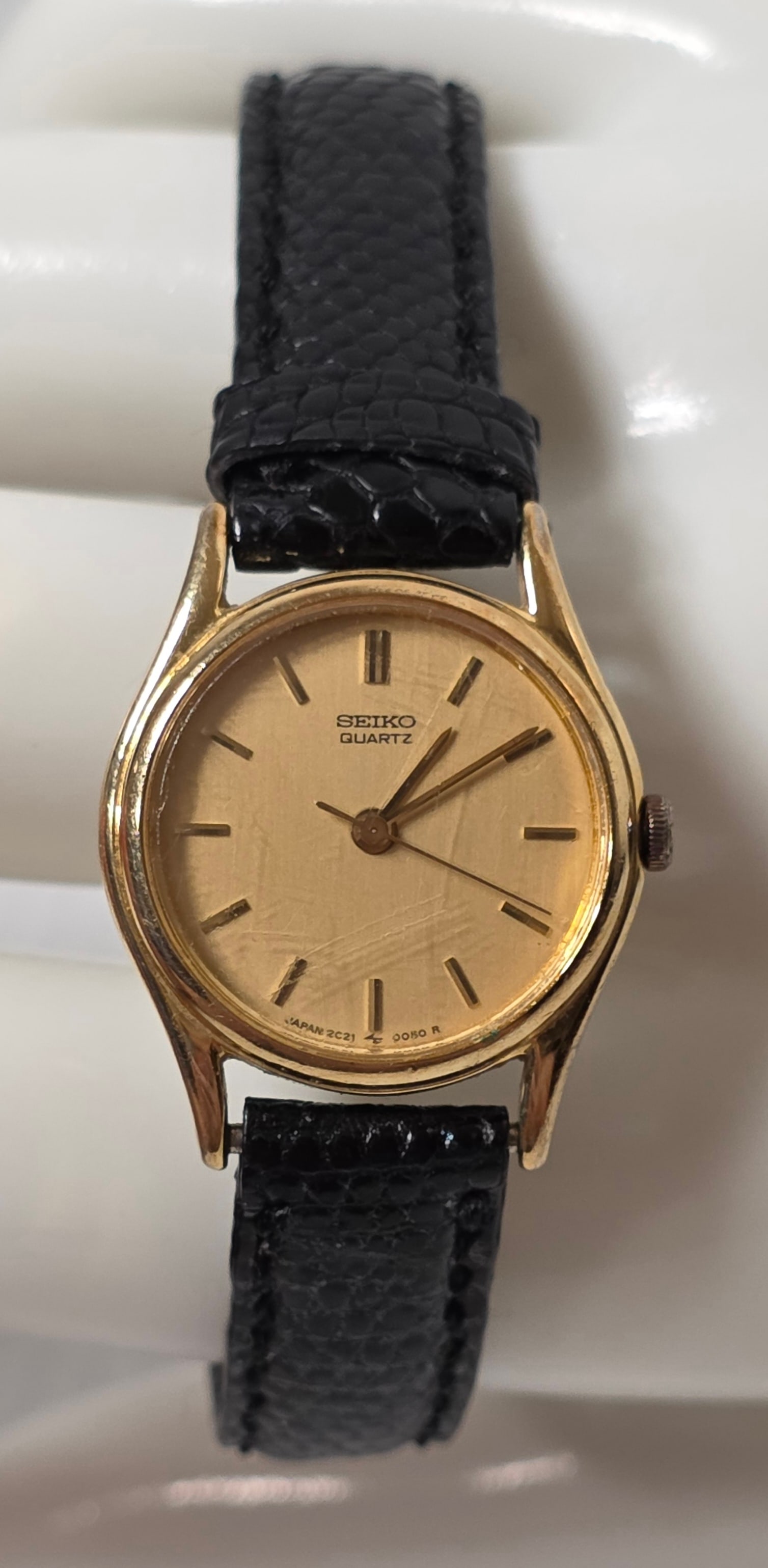Seiko Quartz Gold Tone Ladies Watch Black Strap: Seiko Quartz ladies wristwatch with gold tone case and champagne dial. Features gold tone hour markers and hands with black leather strap. Dial marked Seiko Quartz. Bottom of dial marked Japan 2C21 90