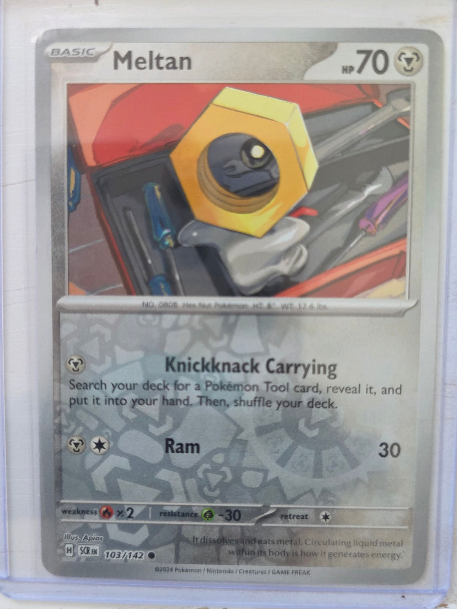 2024 Meltan SCR 103/142 Reverse Holo: 2024 Meltan Basic Metal type Pokemon. SCR set. Card number 103/142. HP 70. Attacks Knickknack Carrying and Ram 30. Reverse holo pattern visible across card surface. Preowned card stored in penny sleev