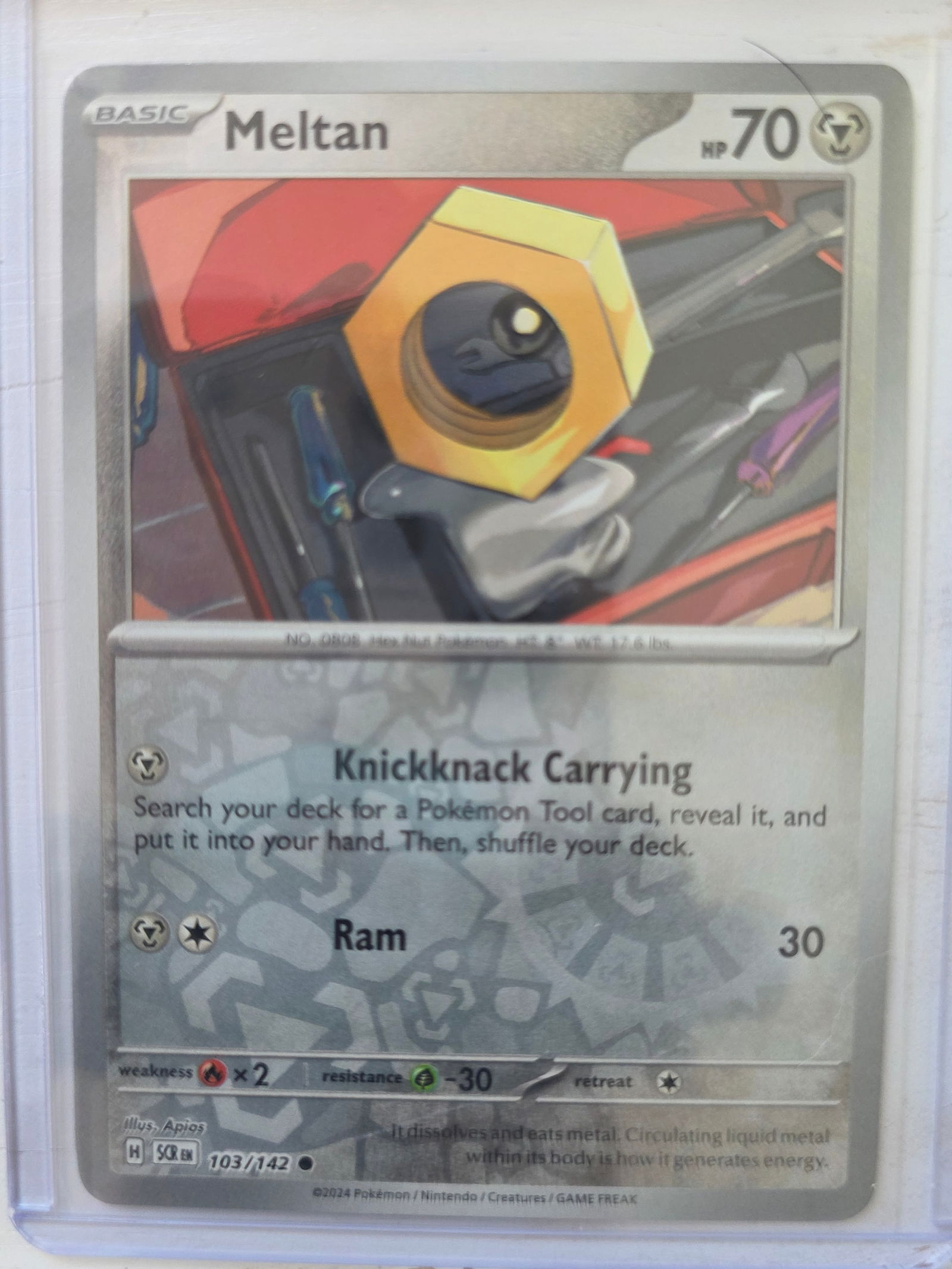 2024 Meltan 103/142 Reverse Holo: 2024 Meltan Basic card 103/142. 70 HP Metal type. Reverse holo foil pattern across card surface with non holo artwork box. Knickknack Carrying ability and Ram 30. Preowned card stored in penny sleeve