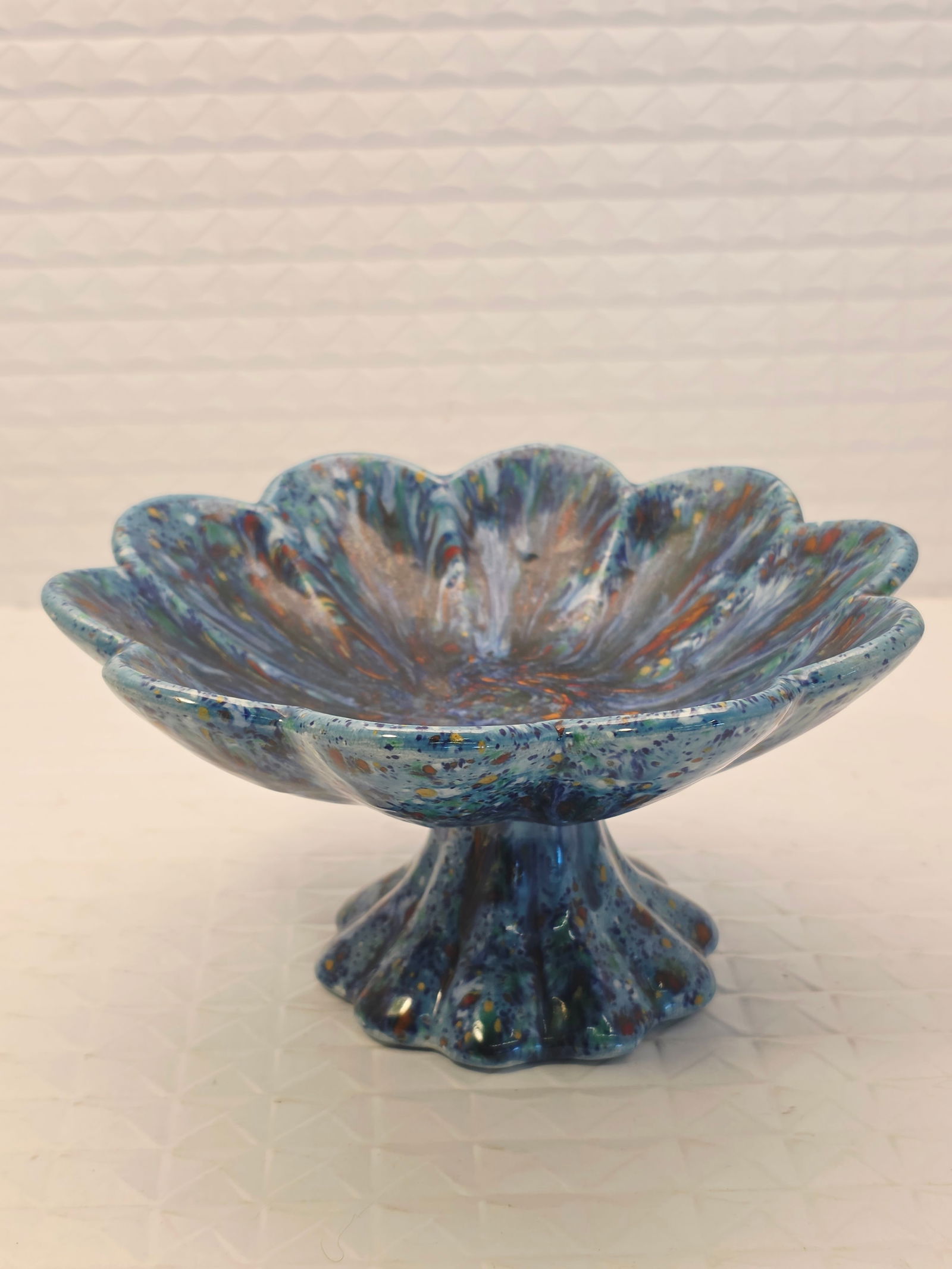 blue multicolored fused glass footed dish: blue multicolored fused glass footed dish or bowl. Material: Fused glass.Style: Features a fluted or scalloped shape with a pedestal base.Purpose: Suitable for use as an accent decorative piece, such