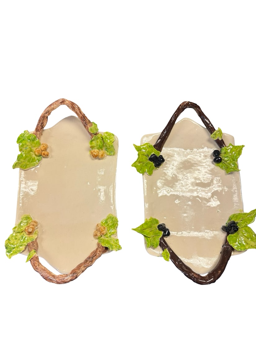 Pair of Botanical Glazed Ceramic Plaques: This pair of handmade ceramic wall plaques features cream-colored backgrounds accented with high-relief sculpted branches. One plaque is decorated with green leaves and light brown acorns, while the o