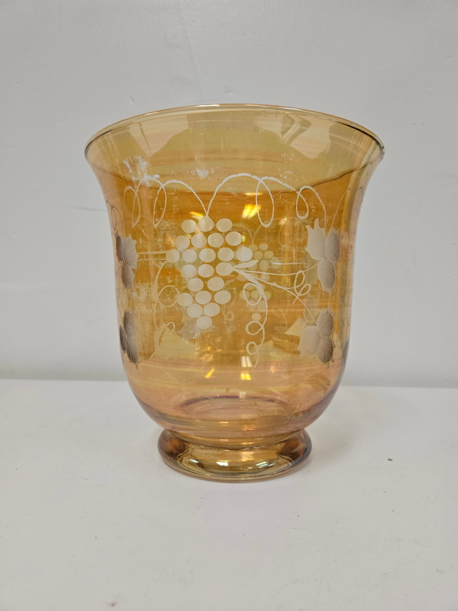 Amber Iridescent Enameled Grape Vase: This large pedestal vase features a warm amber hue with a subtle iridescent carnival glass finish. The exterior is decorated with an enameled white grape and vine motif encircling the flared body.