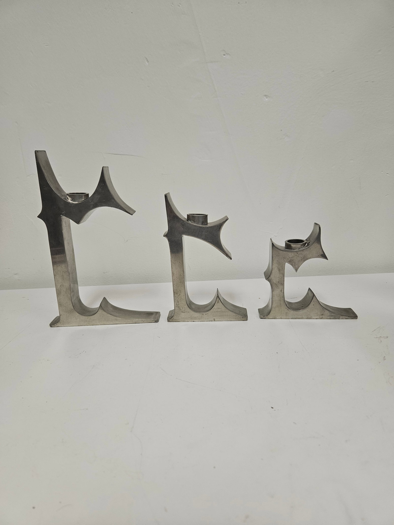 Brutalist Style Graduated Candlesticks: This set of three Brutalist-style candlesticks features a graduated height design with sharp, geometric profiles. Crafted from a heavy cast metal with a matte silver-tone finish, they evoke the indust