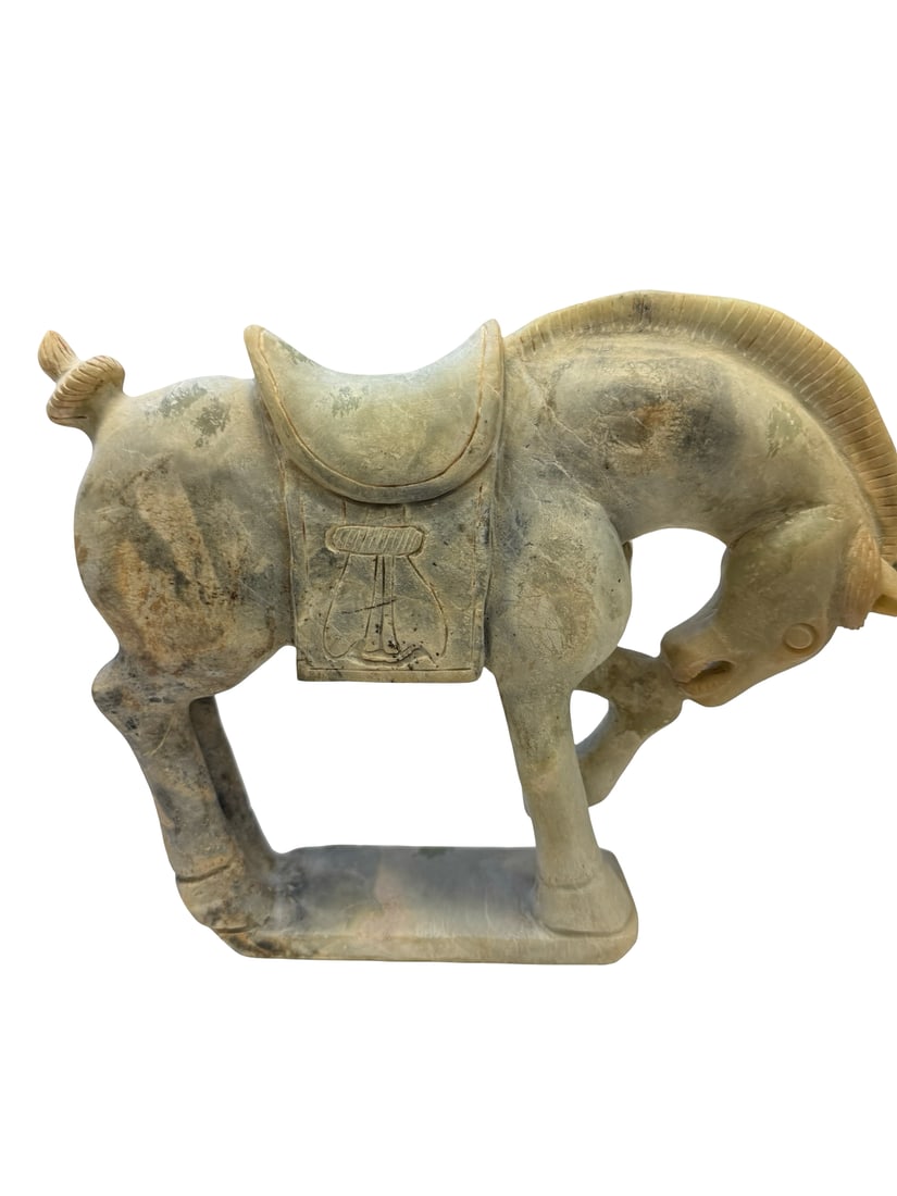 Tang-Style Carved Stone Horse Figure: A carved stone figure depicting a Tang-style caparisoned horse in a dynamic three-legged pose. The sculpture features a detailed saddle, saddlecloth, and cropped mane, crafted from a pale green stone 