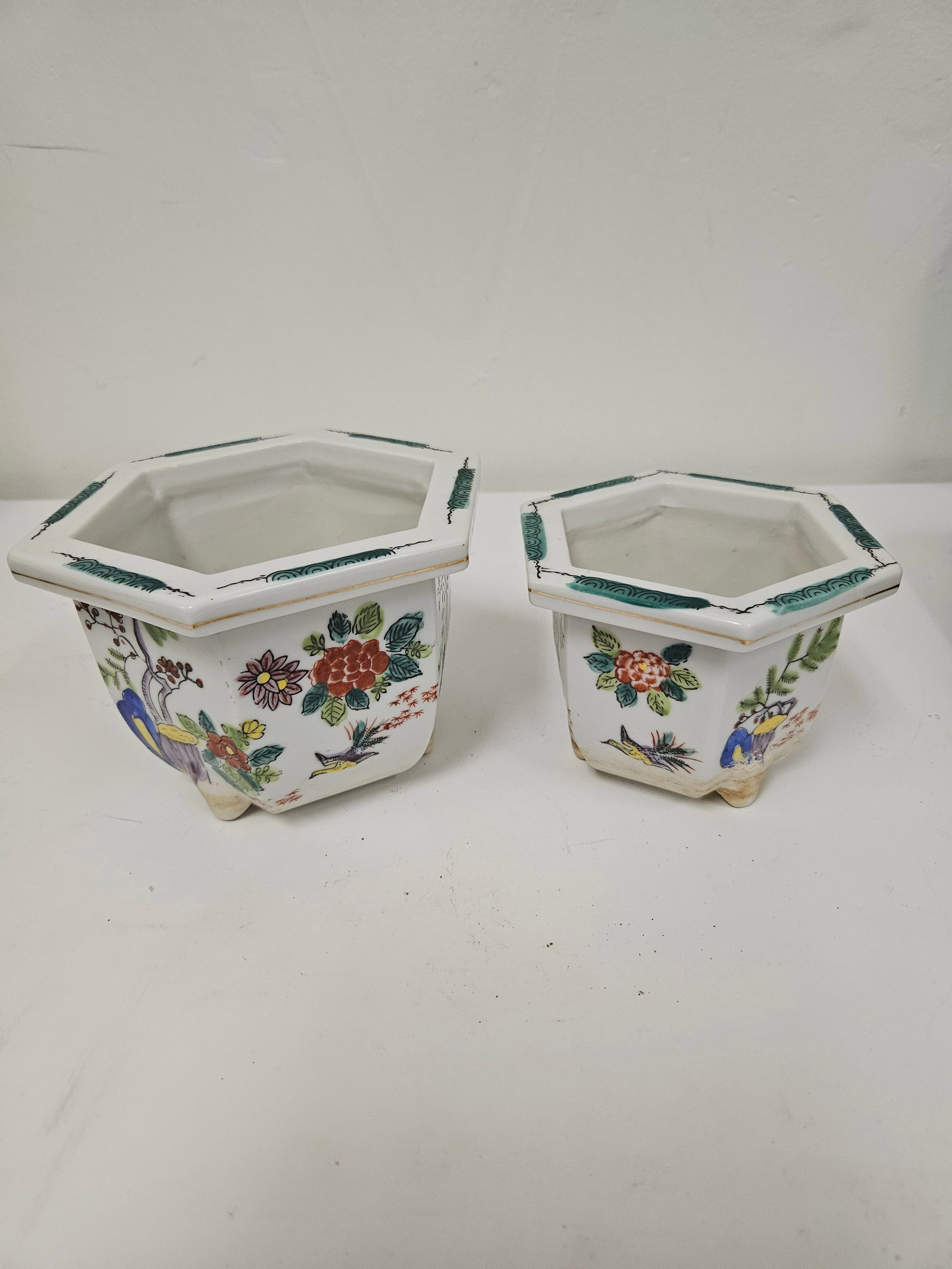 Pair of Hexagonal Floral Ceramic Pots: A pair of graduating hexagonal ceramic planters featuring Famille Rose style floral and bird motifs. Each footed pot includes a central drainage hole and decorative teal scrolling along the rim. The