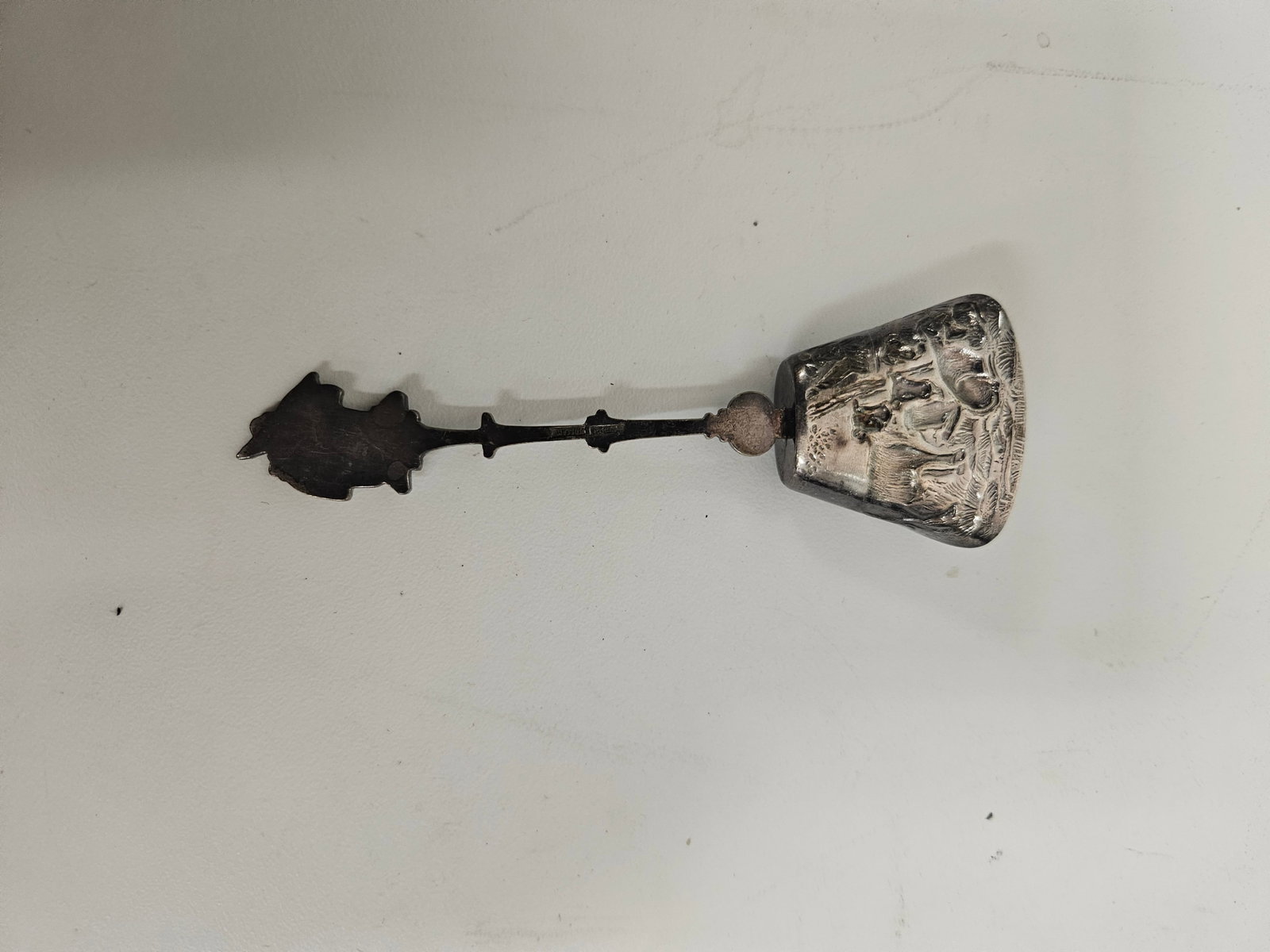 Vintage Ornate Scenic Souvenir Spoon: This vintage metal souvenir spoon features a decorative cast handle ending in a scenic finial. The spade-shaped bowl is embossed with a detailed landscape scene featuring figures and architecture. A d