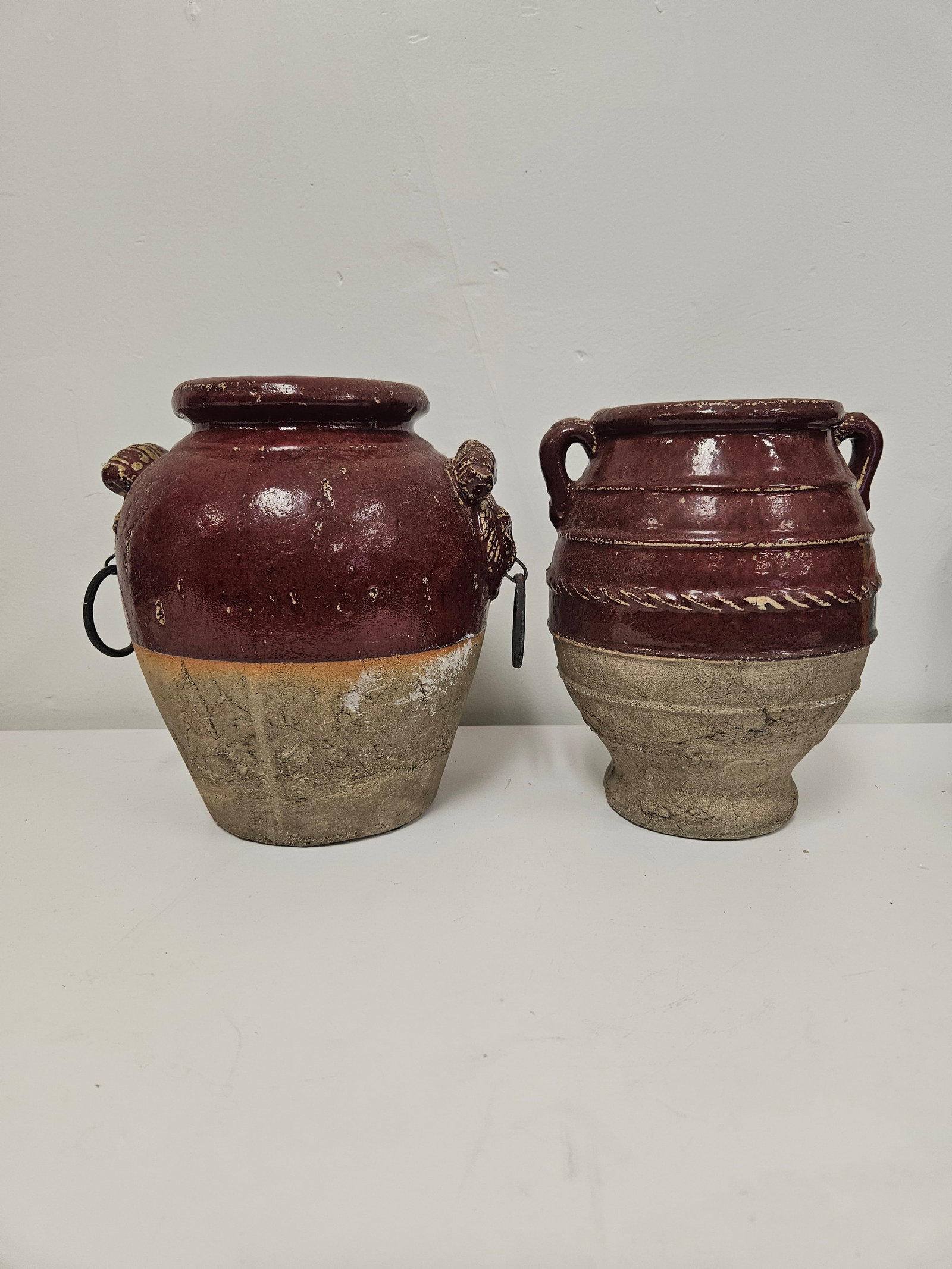 Pair of VG Rustic Two-Tone Ceramic Jars: This pair of decorative ceramic vessels features a rustic two-tone finish with a deep burgundy glaze over a textured, sandy-colored base. One jar includes sculpted lion mask handles with inset metal r
