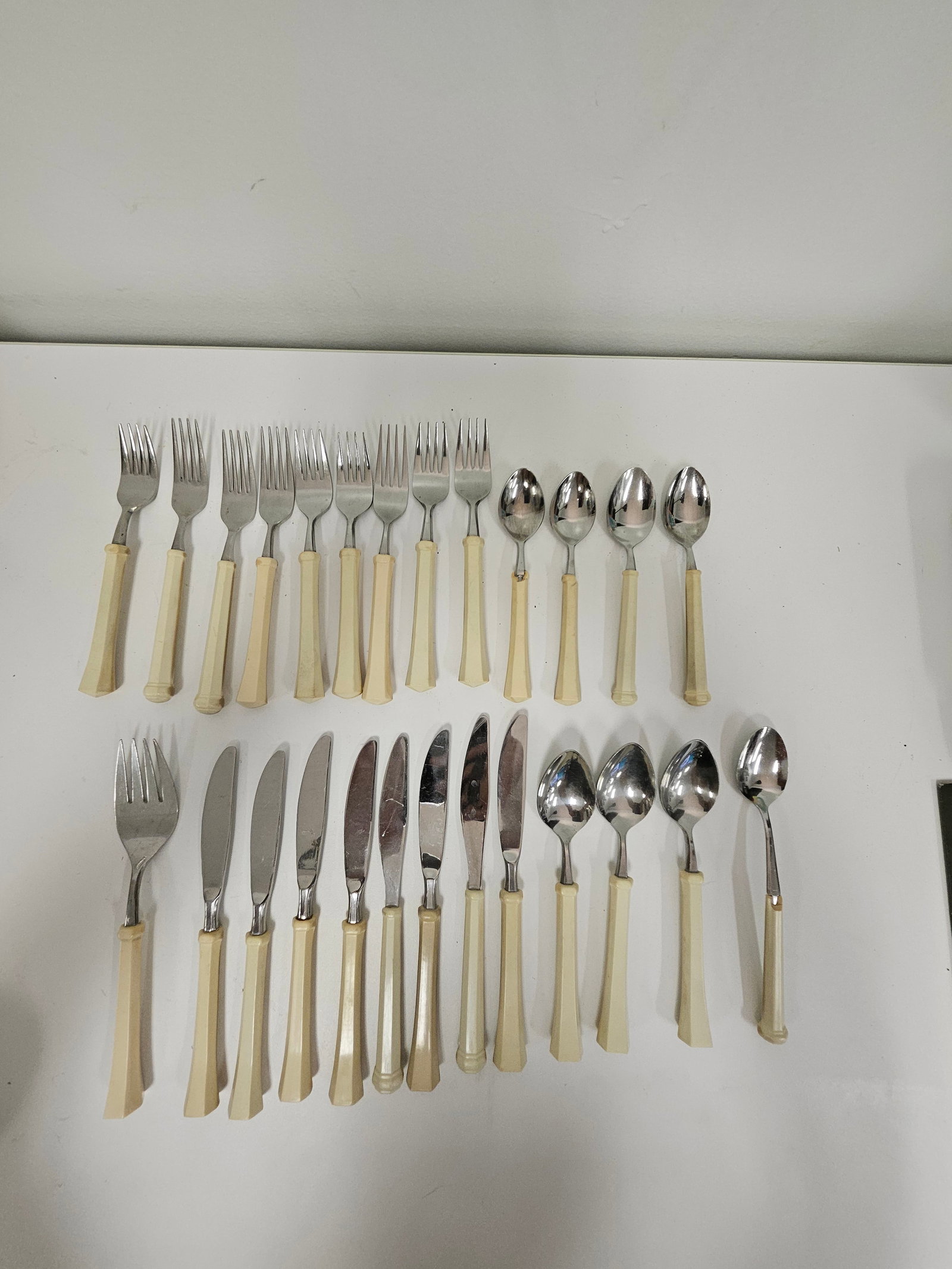 28pc Bakelite Style Handle Flatware Set: This 28-piece flatware set features cream-colored bakelite or early plastic handles with a faceted geometric design. The collection consists of various knives, forks, and spoons made of stainless stee