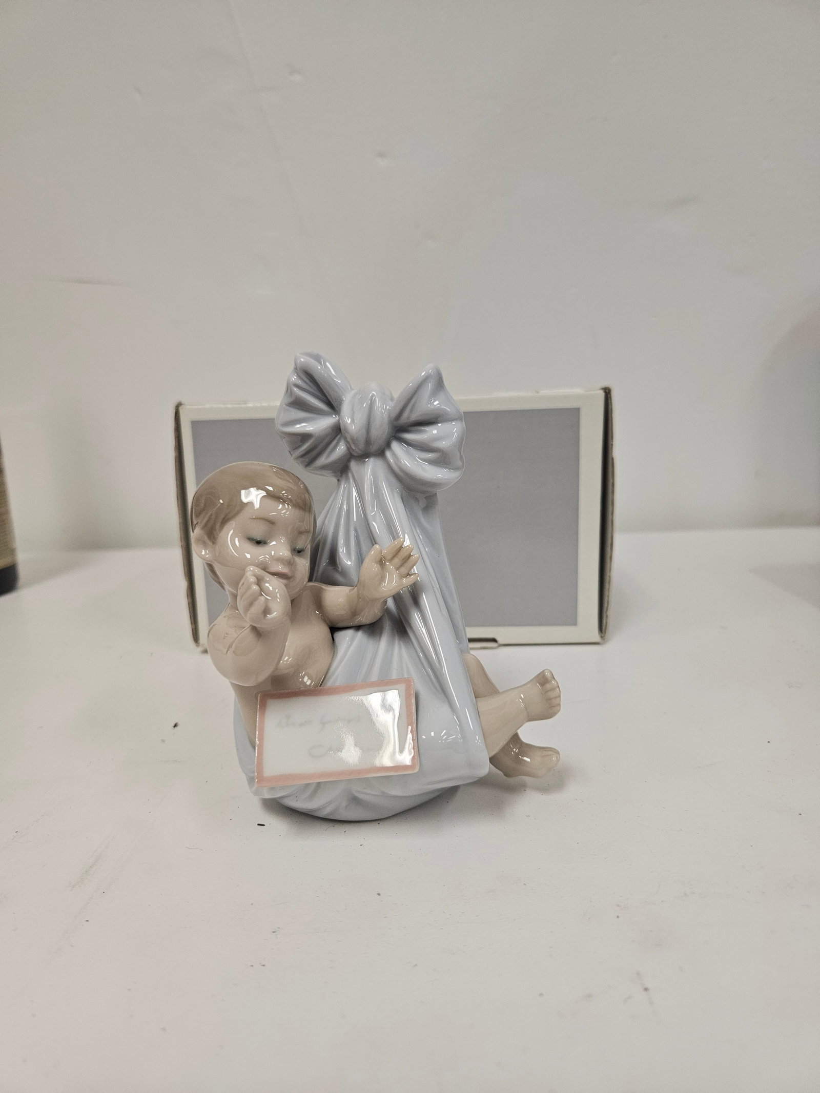 Lladró Newborn Baby Figurine In Box: This porcelain figurine depicts a newborn infant nestled in a blue cloth bundle tied with a large bow. The piece features a glossy finish and soft pastel tones characteristic of Lladró artistry. 