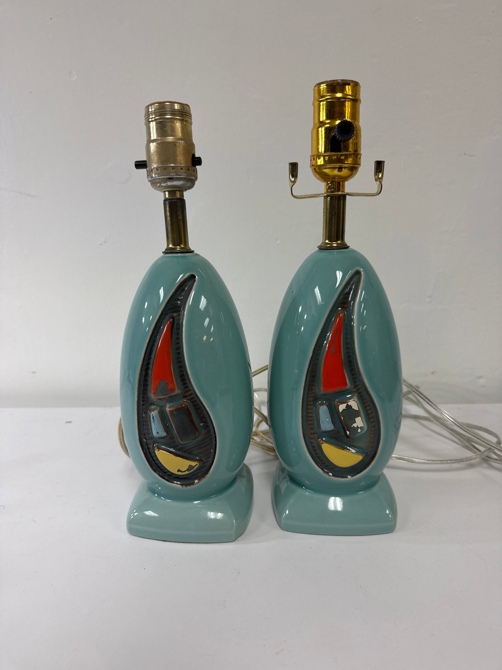 Mid-Century Style Blue Ceramic Lamp Pair: A pair of mid-century modern style ceramic table lamps featuring a distinctive robin's egg blue glaze. Each lamp boasts a teardrop or paisley-shaped cutout on the front housing a colorful geometric ab