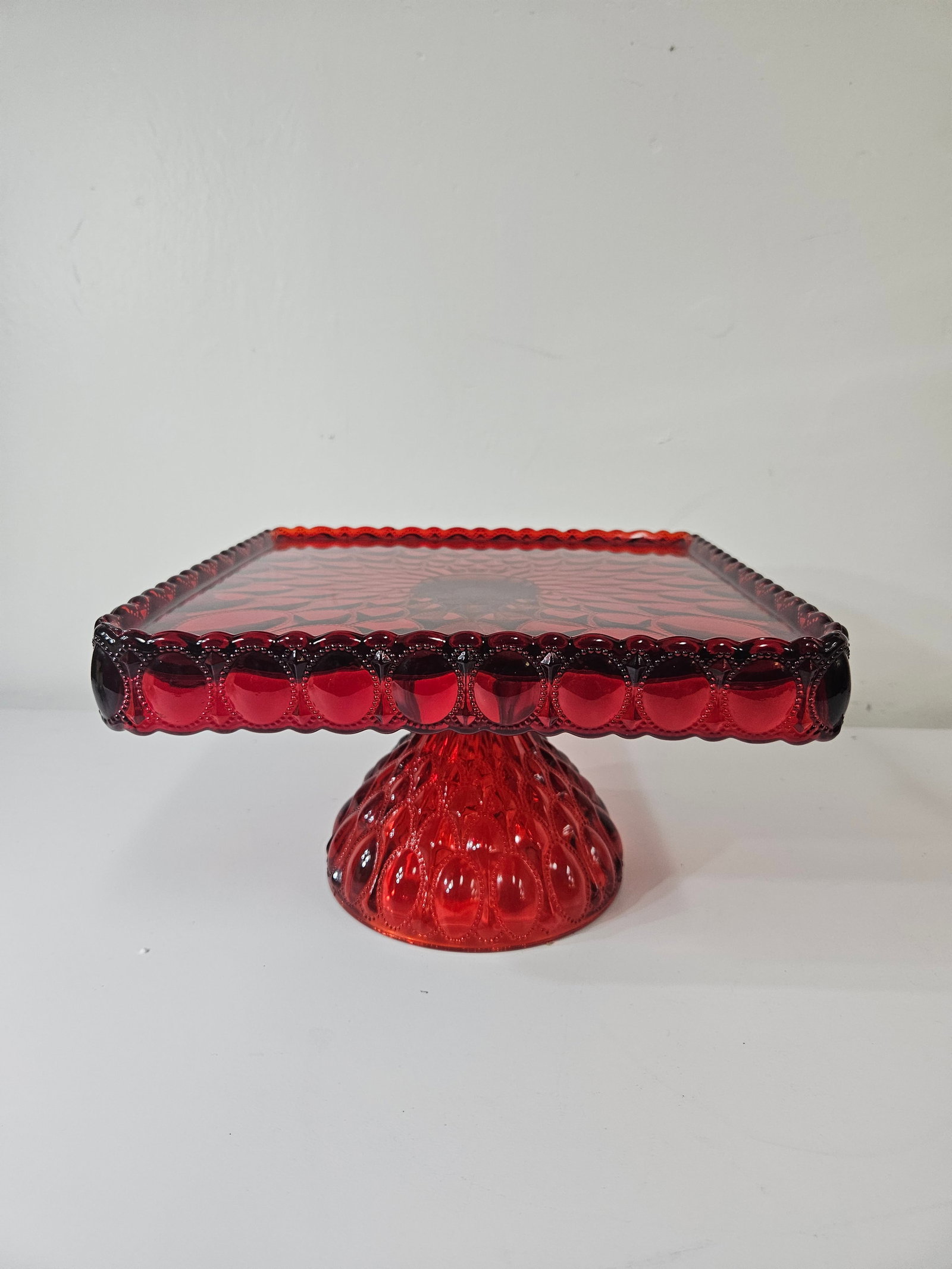 Vintage Ruby Red Thumbprint Cake Stand: This vintage ruby red pressed glass cake stand features a square top with a decorative beaded Thumbprint pattern. The piece is supported by a robust domed pedestal base with matching textured detailin