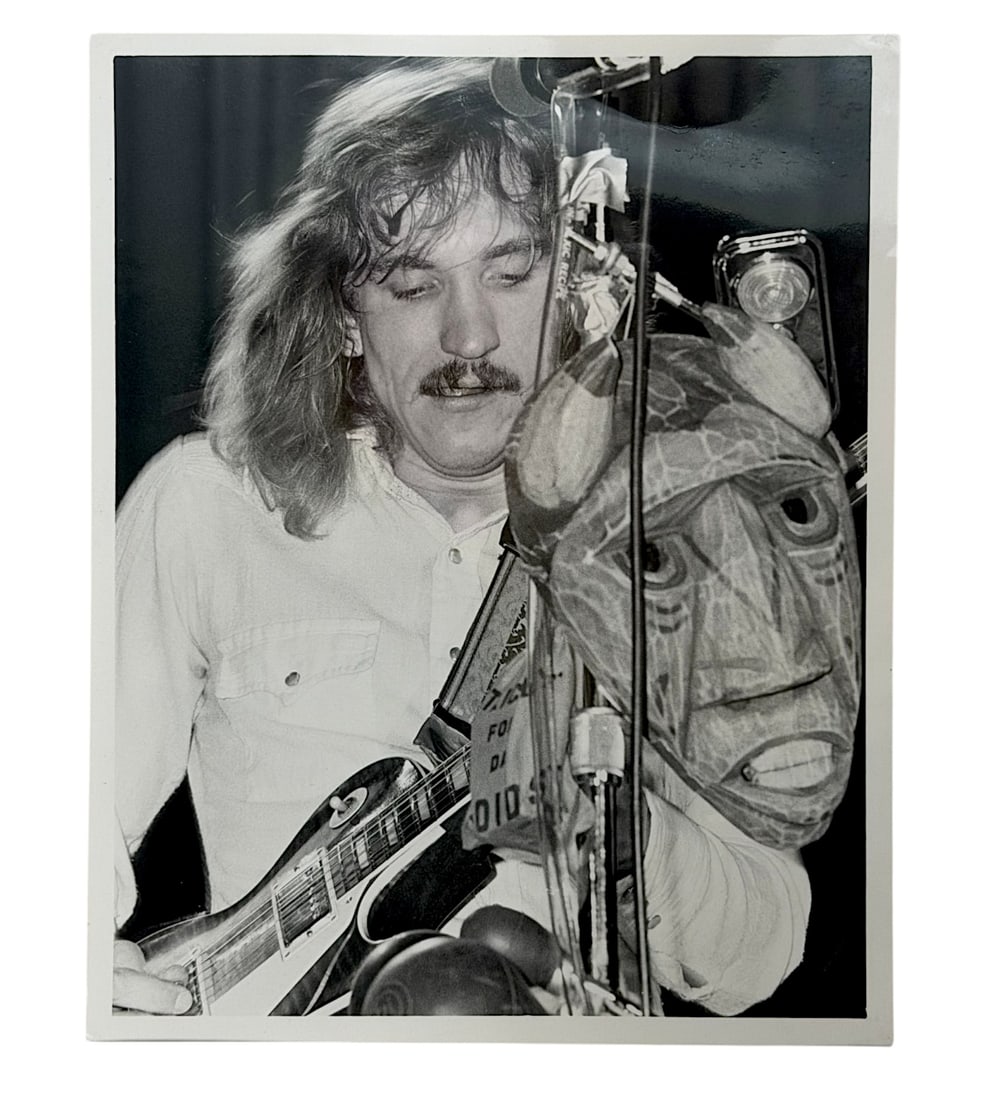 Vintage Rock Music Performance Photograph: Black-and-white concert photograph depicting a guitarist performing on stage alongside a carved mask prop. Vintage music memorabilia photograph.