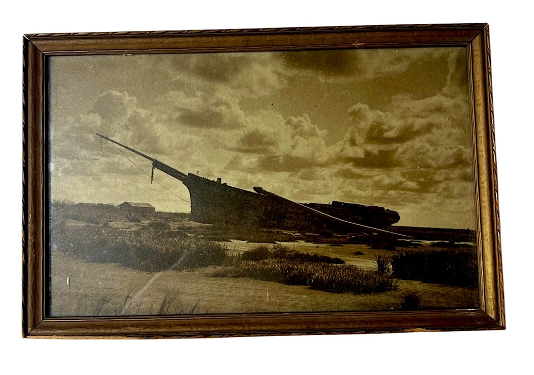 Framed Sepia Shipwreck Photograph: Framed sepia-toned photograph/print of a stranded boat or shipwreck in a coastal landscape. Vintage-style nautical wall decor with frame wear.