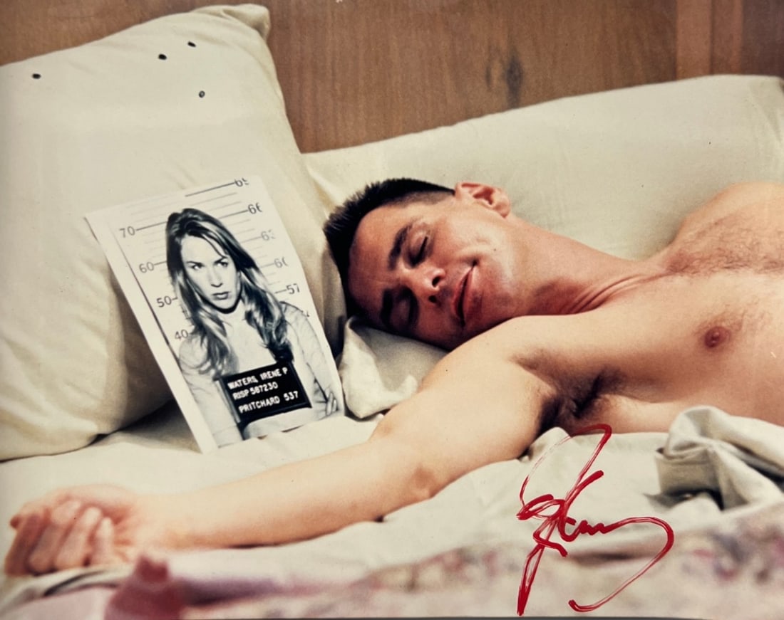 Signed Color Actor Jim Carrey Photograph Bed Scene with Mugshot Me Myself Irene: Color photograph depicting a shirtless male figure reclining in bed beside a printed mugshot-style image of a woman. The photograph bears a large red-ink signature at lower right. The signer and subje