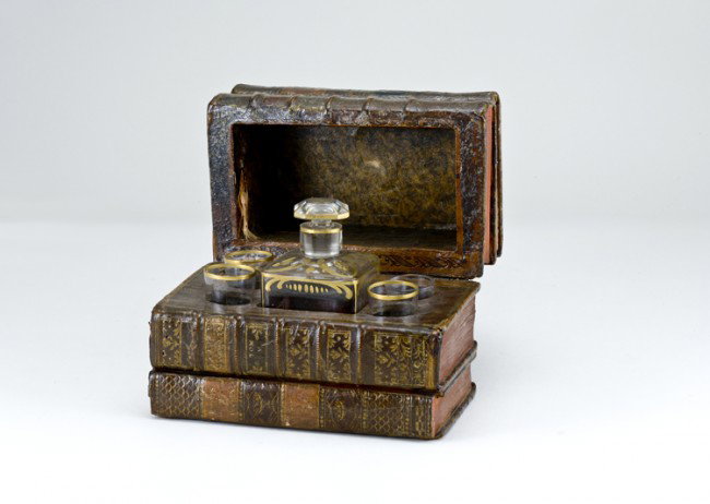 Early Book Box Liquor Set: Measures 6 3/4" x 4" x 4 7/8" tall, interior houses small glass liquor bottle with four shot glasses (one a replacement). Overall in very good condition with wear, may have a later light coat of varni