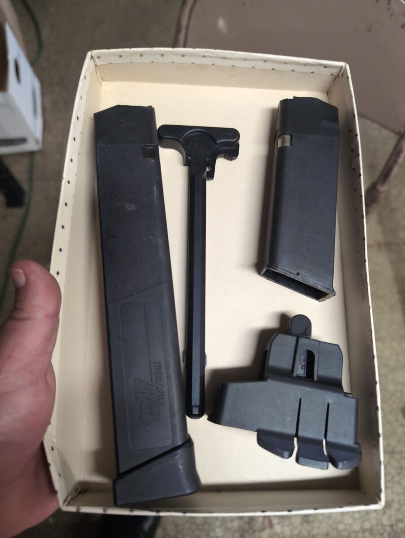 S&M Tactical Firearm Accessory Set: A collection of firearm components including an S&M Tactical extended magazine, a charging handle, a standard magazine, and a polymer accessory mount. Items are black in color and constructed from