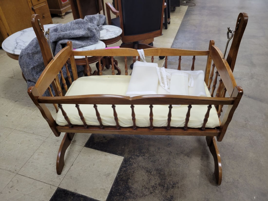 Wooden Spindle Cradle with Mattress and Liner: A wooden cradle featuring a spindle-style design, suspended from a frame. Includes a fitted mattress and a white mesh liner. The unit is designed for rocking motion.