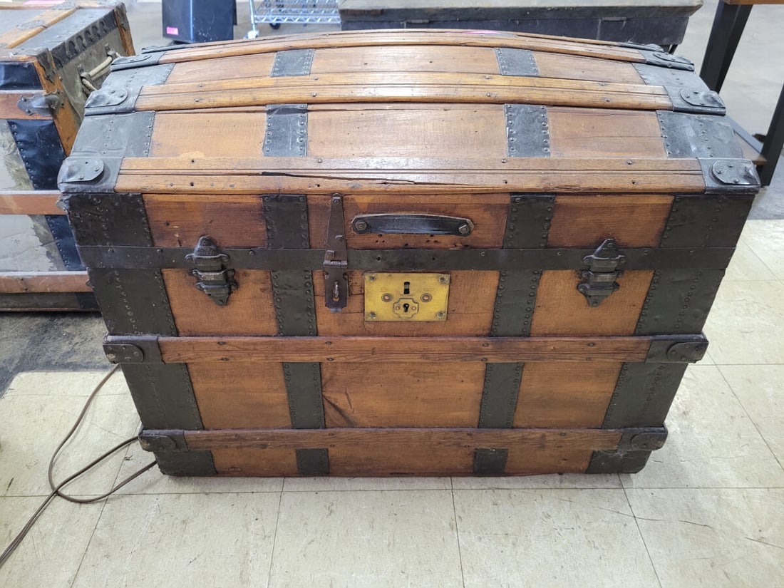 Domed Top Wooden Storage Trunk with Metal Hardware: A wooden storage trunk featuring a domed top design, reinforced with metal bands and corner protectors. The exterior includes side handles and a central brass-toned locking mechanism plate marked