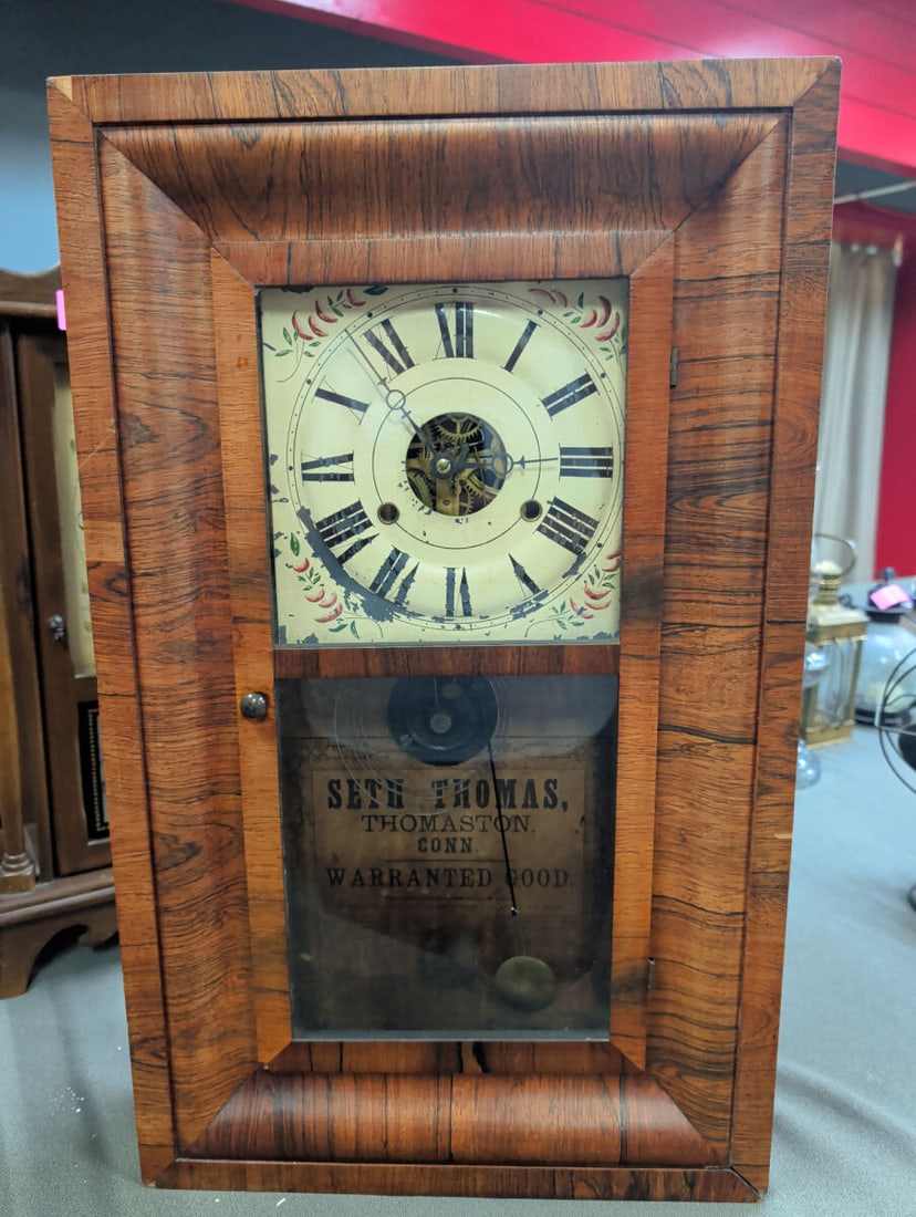 Seth Thomas Thomaston Conn Wall Clock: A wall-mounted clock manufactured by Seth Thomas in Thomaston, Connecticut. The unit features a wooden case with a faux-grain finish, a painted metal dial with Roman numerals and floral accents, and a