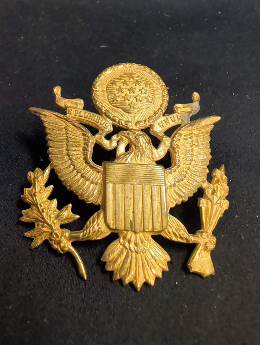 United States Army Officer Cap Insignia: A gold-tone metal United States Army officer cap insignia featuring the Great Seal of the United States. The design depicts a bald eagle with a shield on its chest, clutching an olive branch and a bun