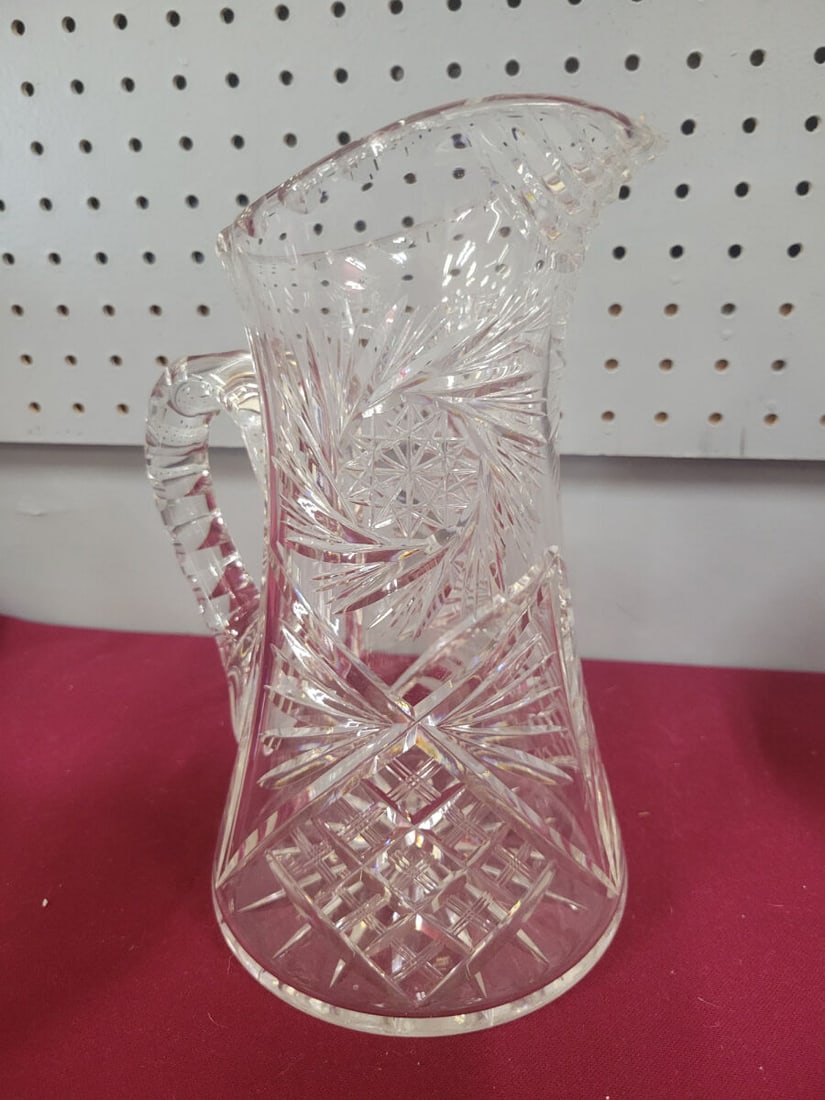Clear Cut Glass Pitcher with Star and Crosshatch P: A clear glass pitcher featuring an intricate cut glass design. The exterior surface is decorated with a repeating pattern of starbursts and crosshatch motifs. The piece includes a sturdy, faceted hand