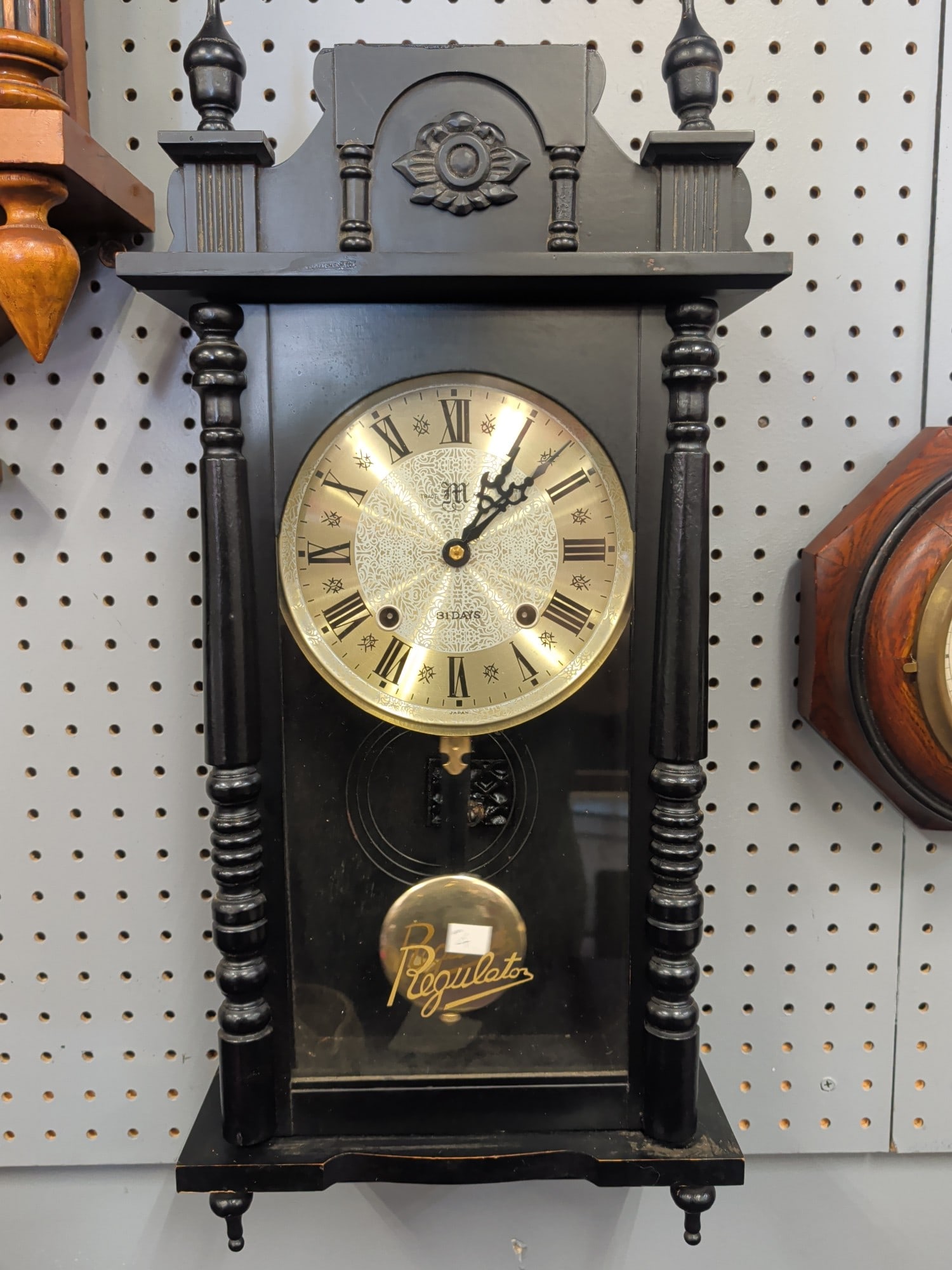 Vintage Japanese Black Regulator Wall Clock with C: Vintage Japanese regulator wall clock featuring a black painted case with turned side columns, decorative crest, brass-tone engraved metal dial with Roman numerals, and lower glazed pendulum compartme