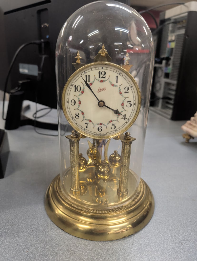Schatz Anniversary Clock with Glass Dome and Flora: German-made Schatz anniversary clock featuring a polished brass case with four decorative columns supporting a white dial accented by delicate floral garlands. The clock is housed beneath its