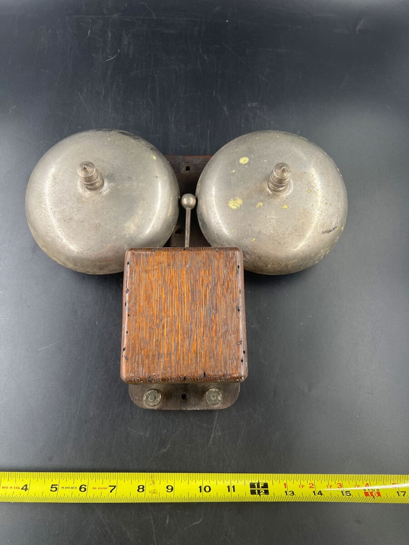 Double Dome Wood Back Electric Call Bell Ringer Mechanism: Electric call bell system featuring a wooden base board, a central covered mechanism housing, dual metal bells on either side, and a central striking clapper. The back mounting board includes pre-dril