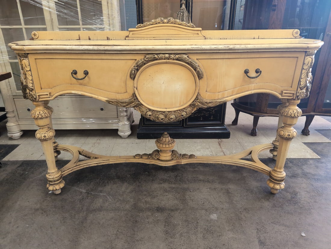 French Country Sideboard: Elaborately carved wood sideboard featuring a cream-colored painted finish with distressed gold-toned accents, floral and scroll motif carvings, two drawers with drop handles, shaped lower stretcher