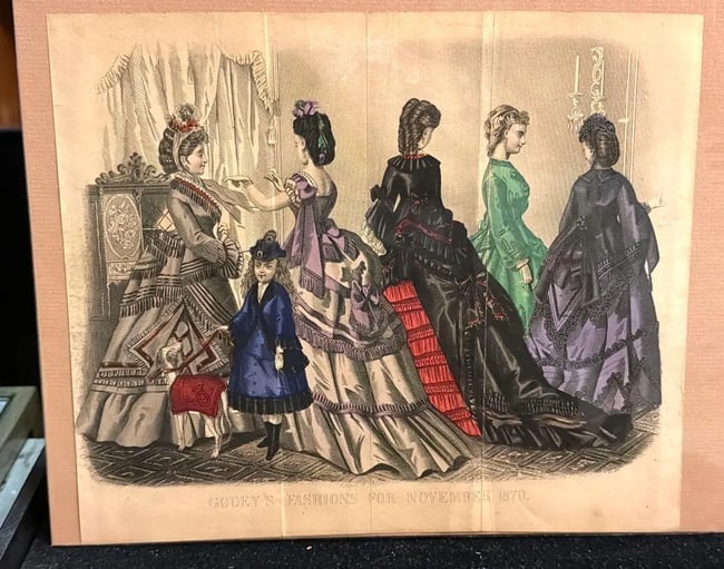 Old Hand Tinted Page from Godey's Fashion for November 1870: A hand-colored fashion plate titled "GODEY'S FASHIONS FOR NOVEMBER 1870". It depicts five women and one child showcasing elaborate Victorian-era clothing silhouettes featuring bustles and elaborate