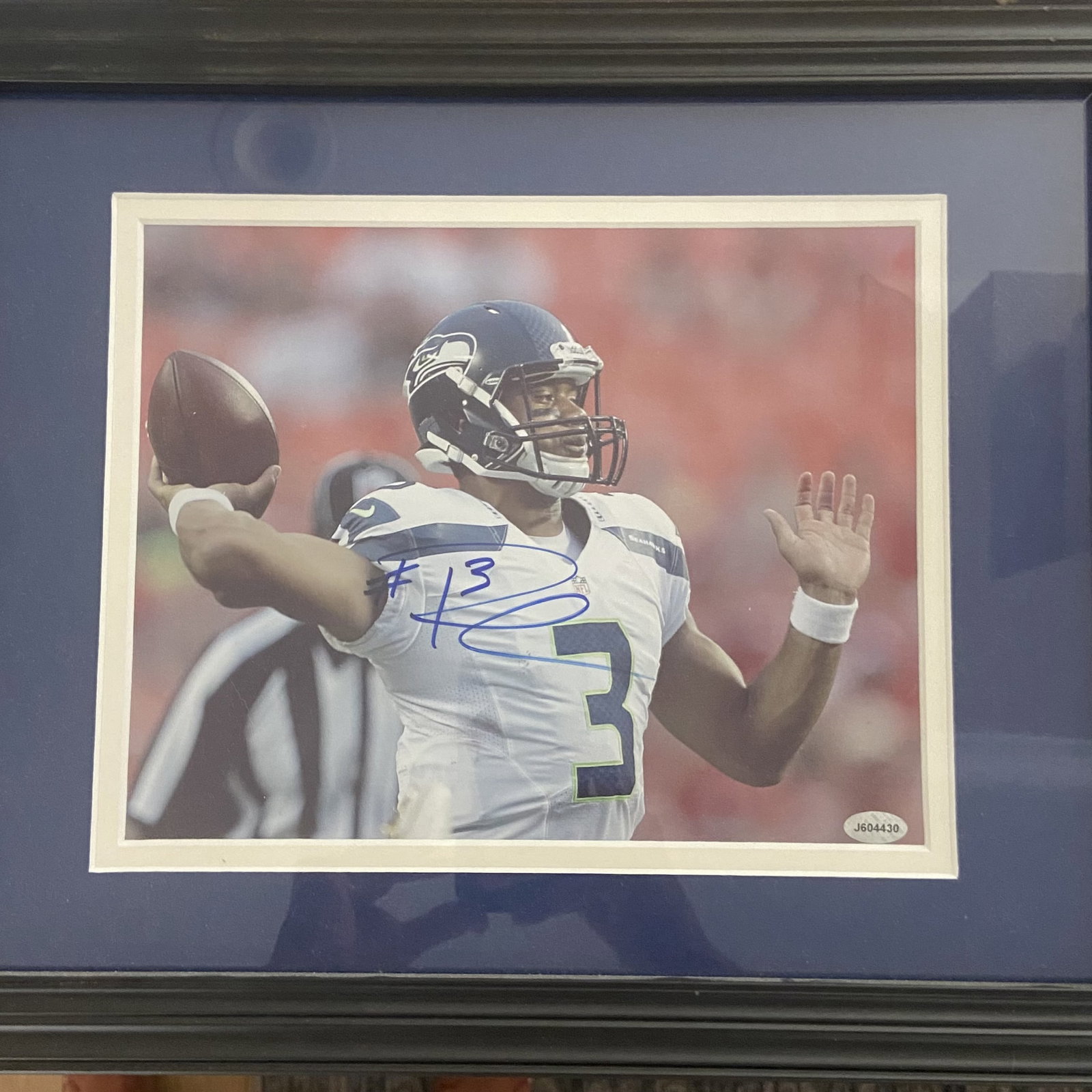 Russell Wilson Autographed Seattle Seahawks Photo: a framed, autographed photograph of Russell Wilson playing for the Seattle Seahawks. The image features Wilson in his white jersey passing the football, accompanied by a JSA certification sticker in t