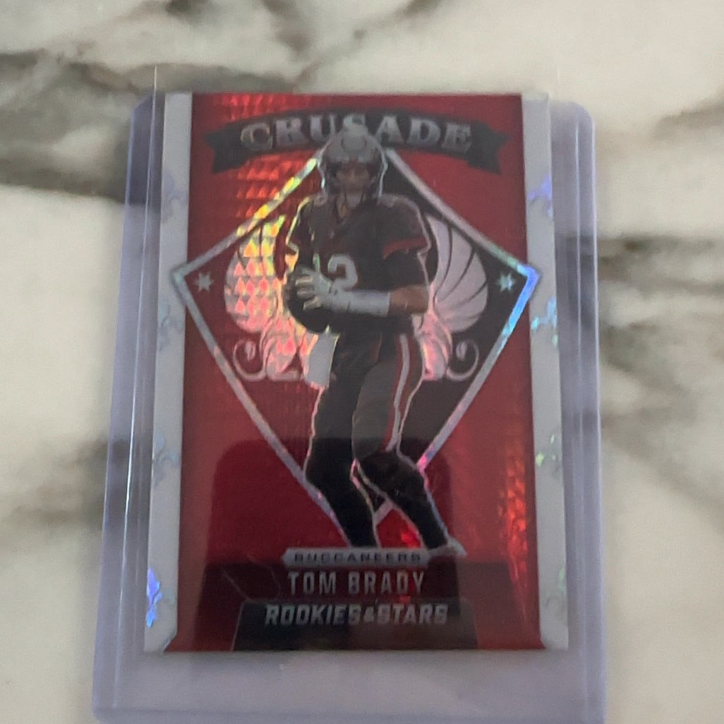 2021 Panini Rookies & Stars Crusade Tom Brady: This is a 2021 Panini Rookies & Stars Crusade Tom Brady football card. It features a red parallel design with Brady in his Tampa Bay Buccaneers uniform.