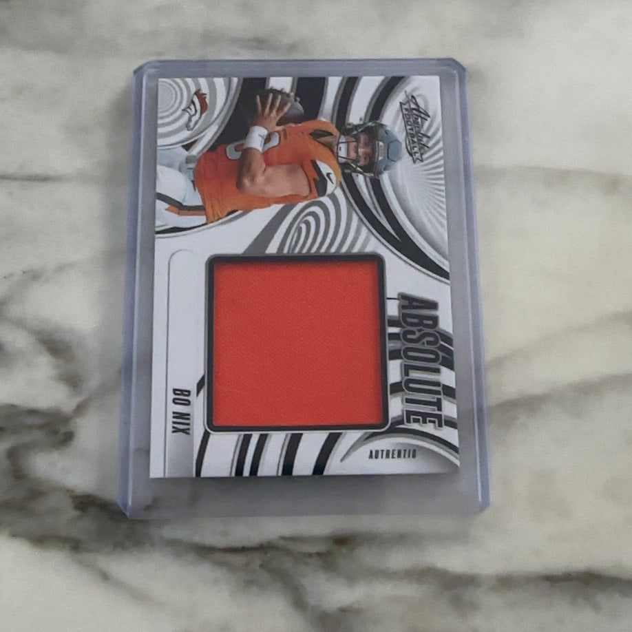 Bo Nix Absolute Football Memorabilia Card: This is a 2024 Panini Absolute Football card featuring a player image of Bo Nix. The card includes a prominent orange fabric relic piece embedded in the front design. It is presented in a protective