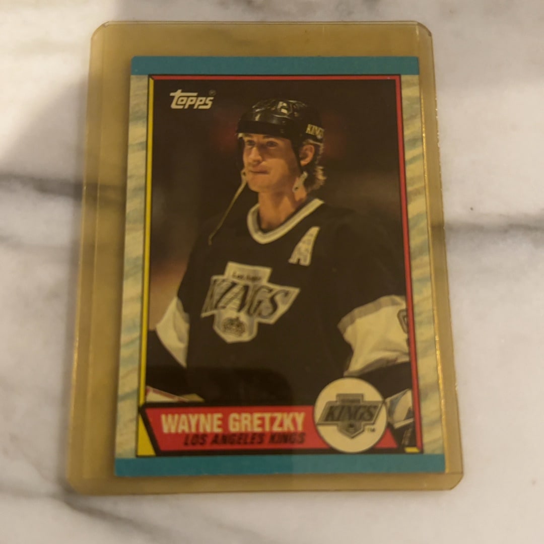 1990 Topps Wayne Gretzky Los Angeles Kings Hockey Card: This is a 1990 Topps hockey card featuring Wayne Gretzky of the Los Angeles Kings. The card displays Gretzky in his iconic black uniform and comes preserved in a protective plastic holder.