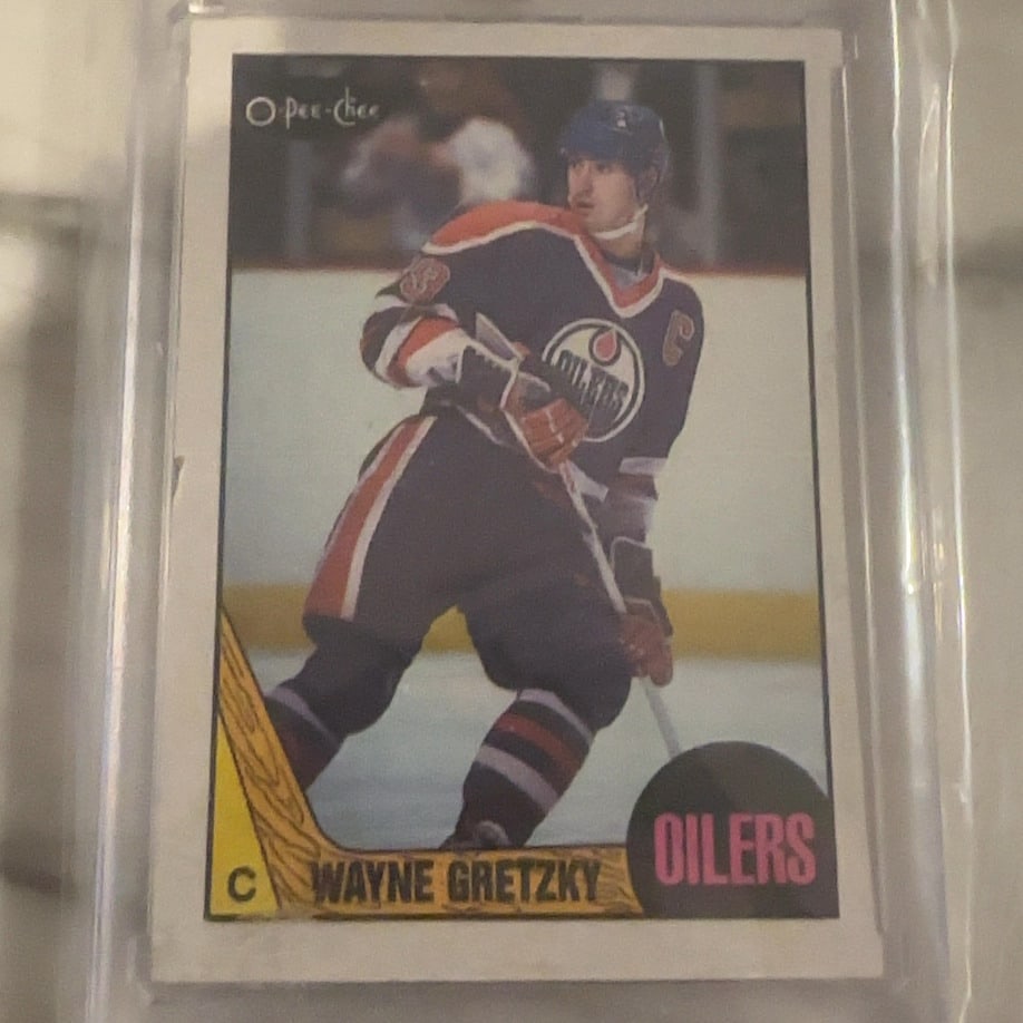 1980 O-Pee-Chee Wayne Gretzky Hockey Card: This is an original 1980 O-Pee-Chee hockey trading card featuring Wayne Gretzky of the Edmonton Oilers. The card displays the iconic wood-grain design characteristic of this set. It is protected withi