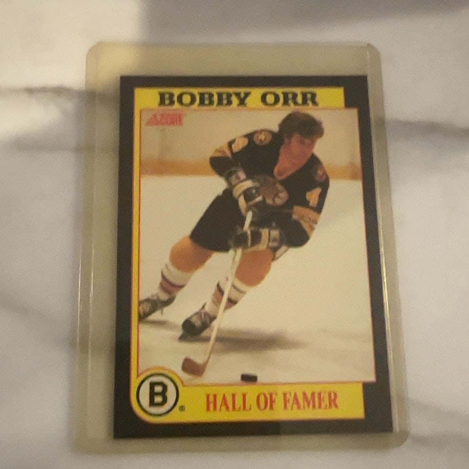 1991 Score Bobby Orr Hockey Card: This is a 1991 Score hockey trading card featuring the legendary Boston Bruins defenseman Bobby Orr. The card identifies him as a Hall of Famer and is preserved in a protective plastic top loader.