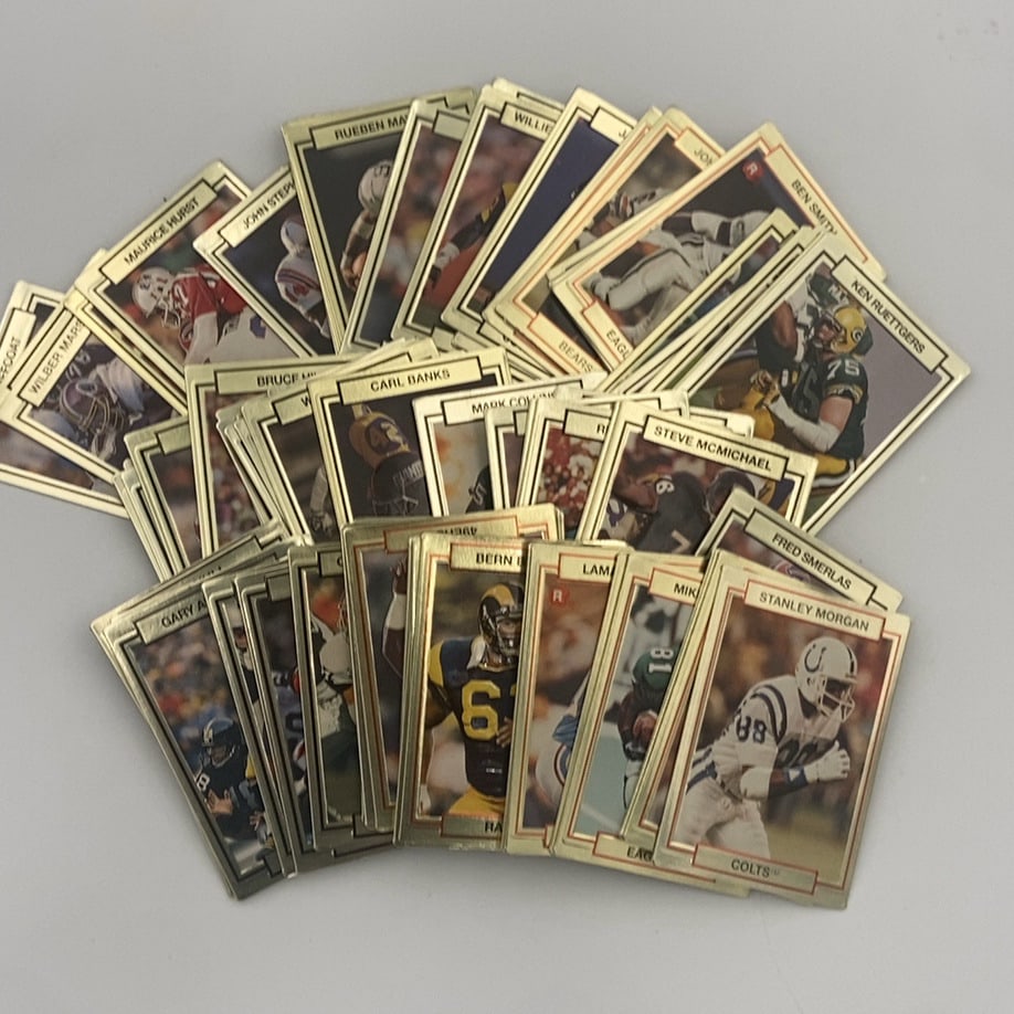 1990 Pro Set NFL Football Trading Card Collection: This is a collection of various 1990 Pro Set NFL football trading cards fanned out. Each card features a gold-colored border design with a player photograph, team helmet, and nameplate at the top,
