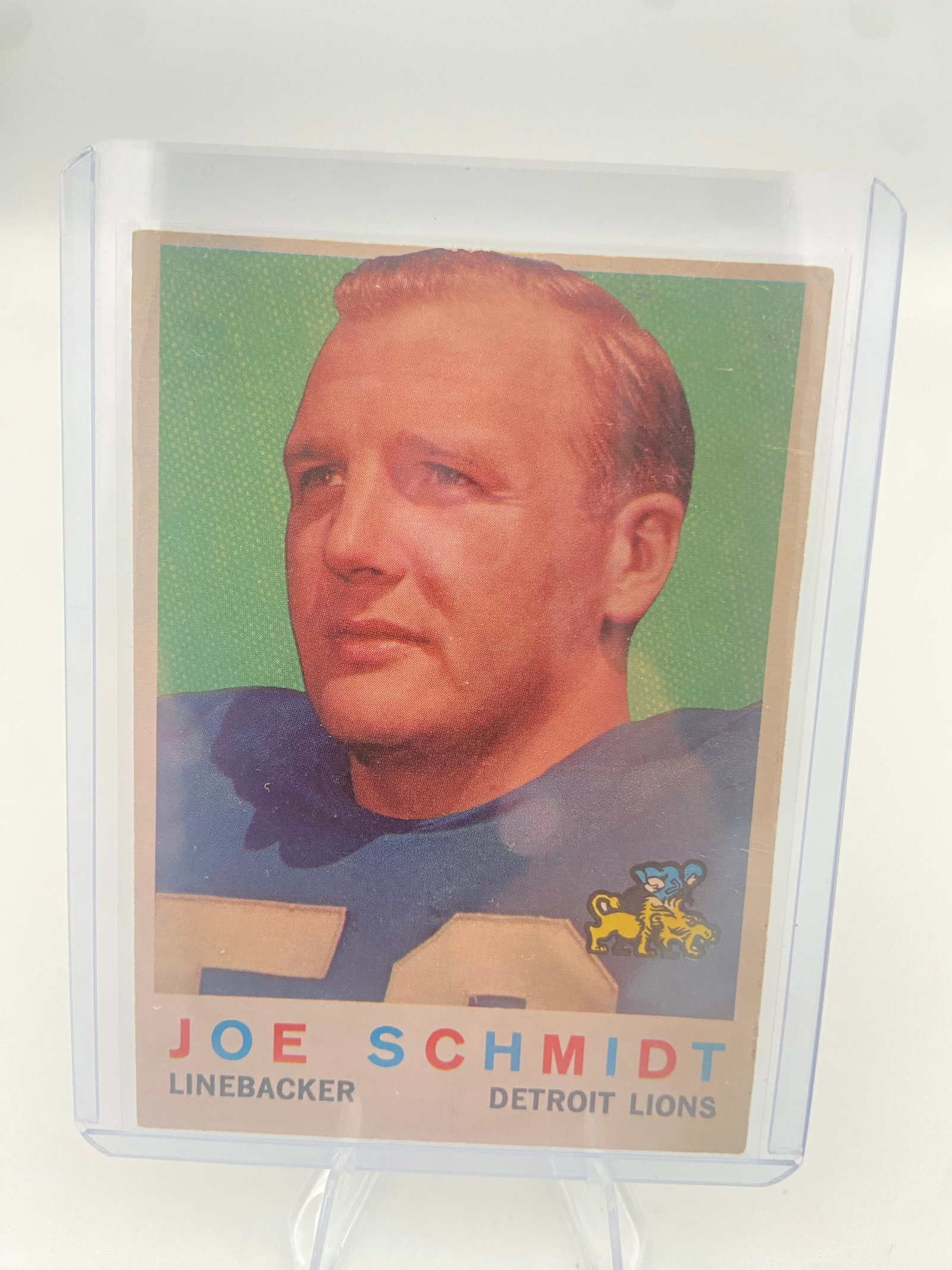 1958 Topps Joe Schmidt Detroit Lions Football Card: This is a 1958 Topps football card featuring Joe Schmidt of the Detroit Lions. The card captures a close-up portrait of the legendary linebacker and is preserved in a protective plastic top-loader.