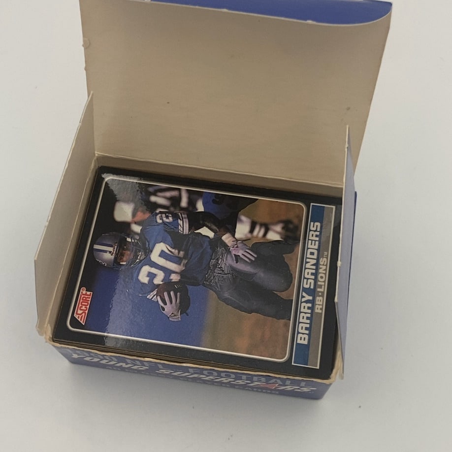 1990 Score Young Superstars Football Card Set: This is a factory sealed box set of 1990 Score Young Superstars football cards, featuring a prominent card of Hall of Fame running back Barry Sanders on the front. The set contains a collection of