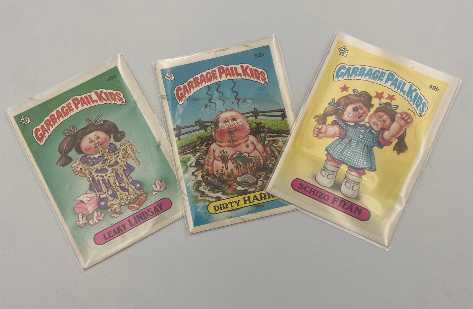 Vintage Garbage Pail Kids Trading Cards: A set of three vintage Garbage Pail Kids trading card stickers including characters Leaky Lindsay, Dirty Harry, and Schizo Fran. These iconic 1980s collectibles feature humorous and grotesque