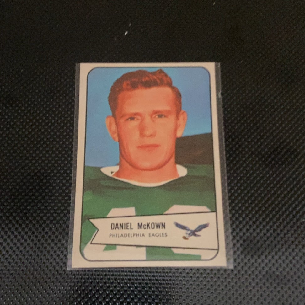 1960 Topps Daniel McKown Philadelphia Eagles Football Card: This is a 1960 Topps football card featuring Philadelphia Eagles player Daniel McKown. The card is preserved in a protective sleeve and depicts a portrait of the athlete against a blue background
